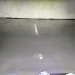 USŁUGI OGÓLNOBUDOWLANE Michał Romanowski - Świeżo wylana, gładka, szara posadzka w narożniku pomieszczenia, z widocznym fragmentem listwy przypodłogowej i odblaskami światła na powierzchni.