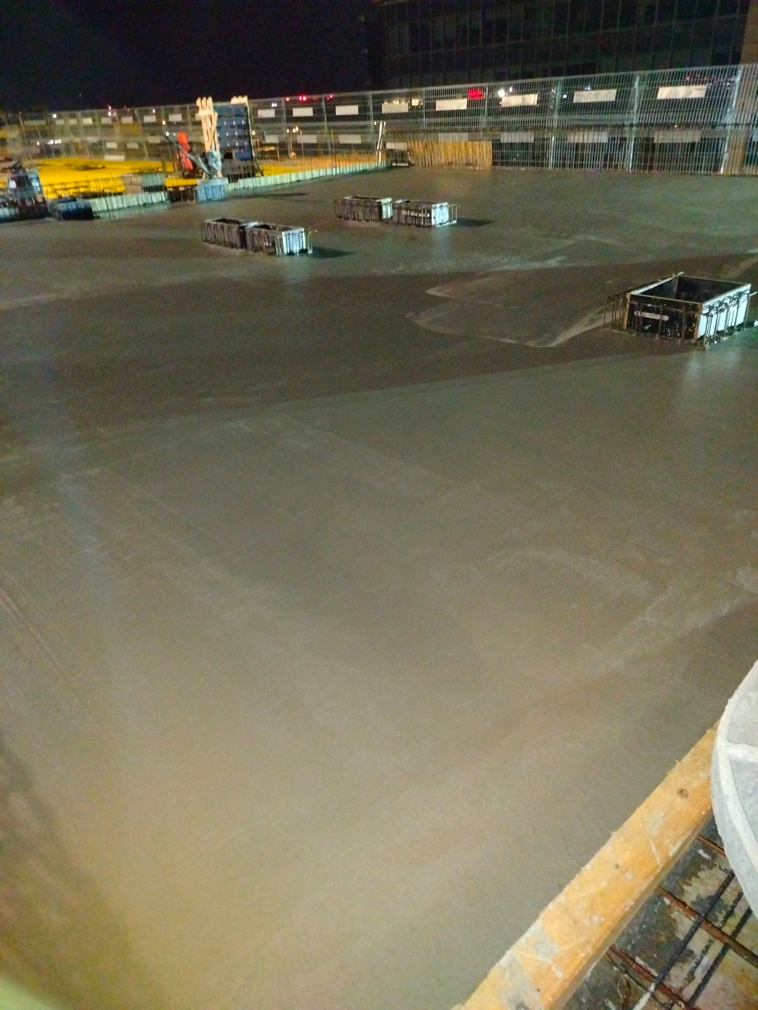 Świeżo wylana, gładka betonowa posadzka przemysłowa w hali produkcyjnej, widoczne metalowe formy i elementy szalunkowe, oświetlenie hali w tle.