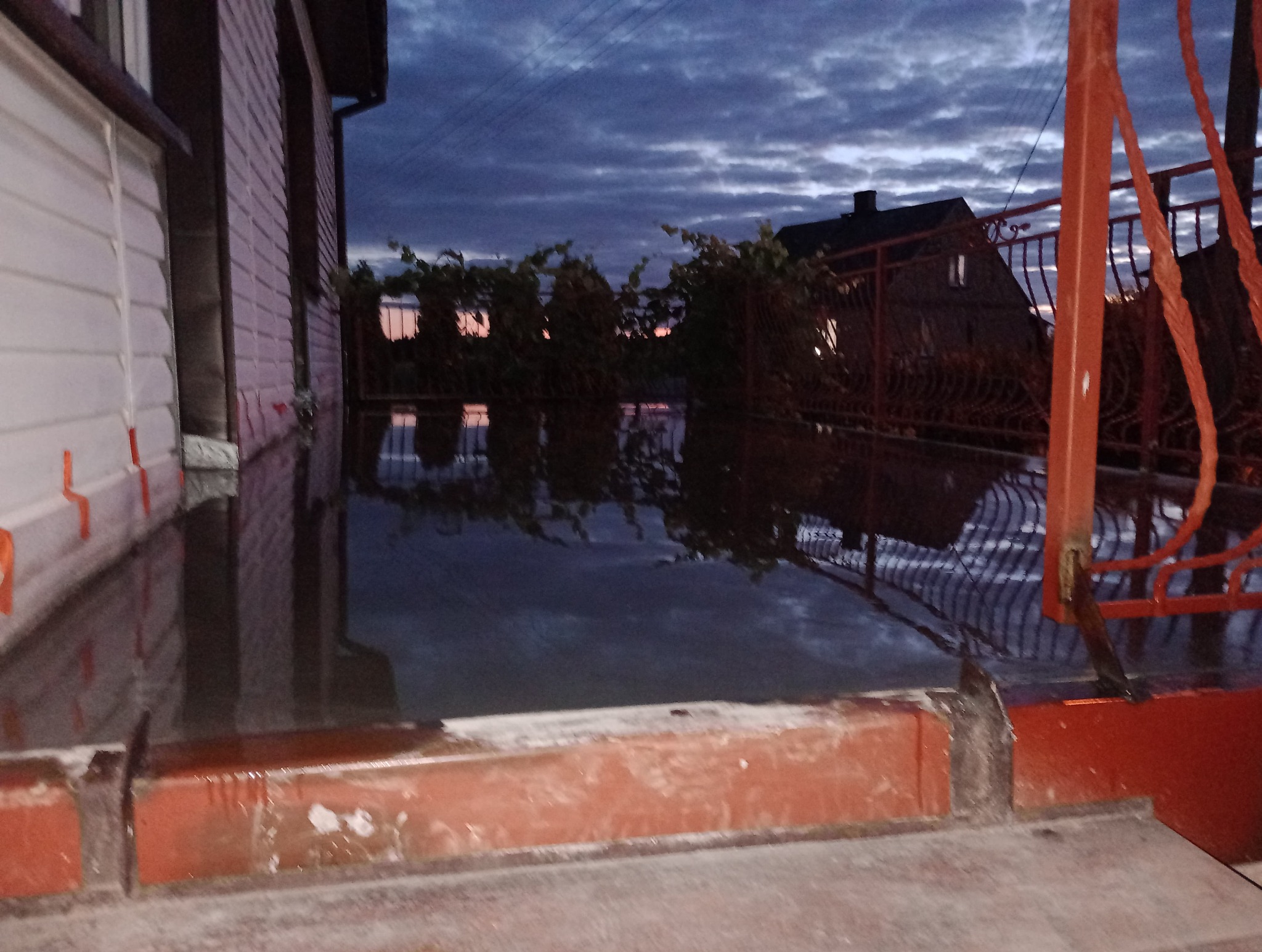 Zalany teren posesji z widocznymi elementami domu murowanego i metalowego ogrodzenia w pochmurny wieczór, odbicie nieba w wodzie.