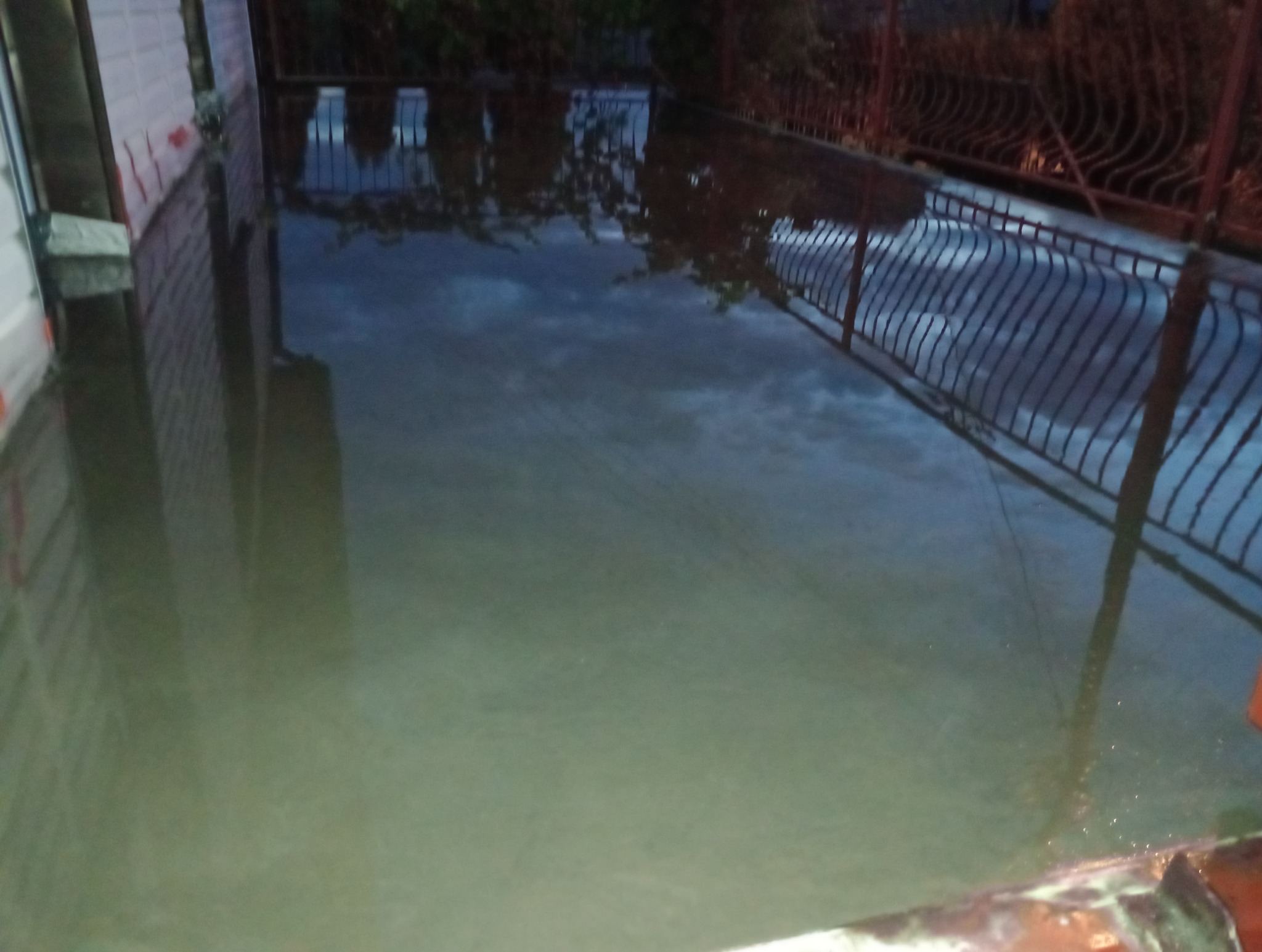 Zalana posadzka na zewnątrz budynku, z odbiciem nieba i metalowego ogrodzenia w wodzie.