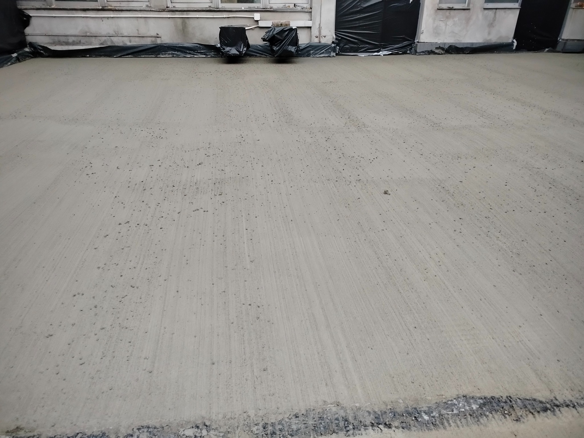 Świeżo wylana, jasnoszara posadzka przemysłowa z widocznymi śladami po narzędziach do rozprowadzania betonu, zabezpieczona czarną folią przy ścianie budynku.