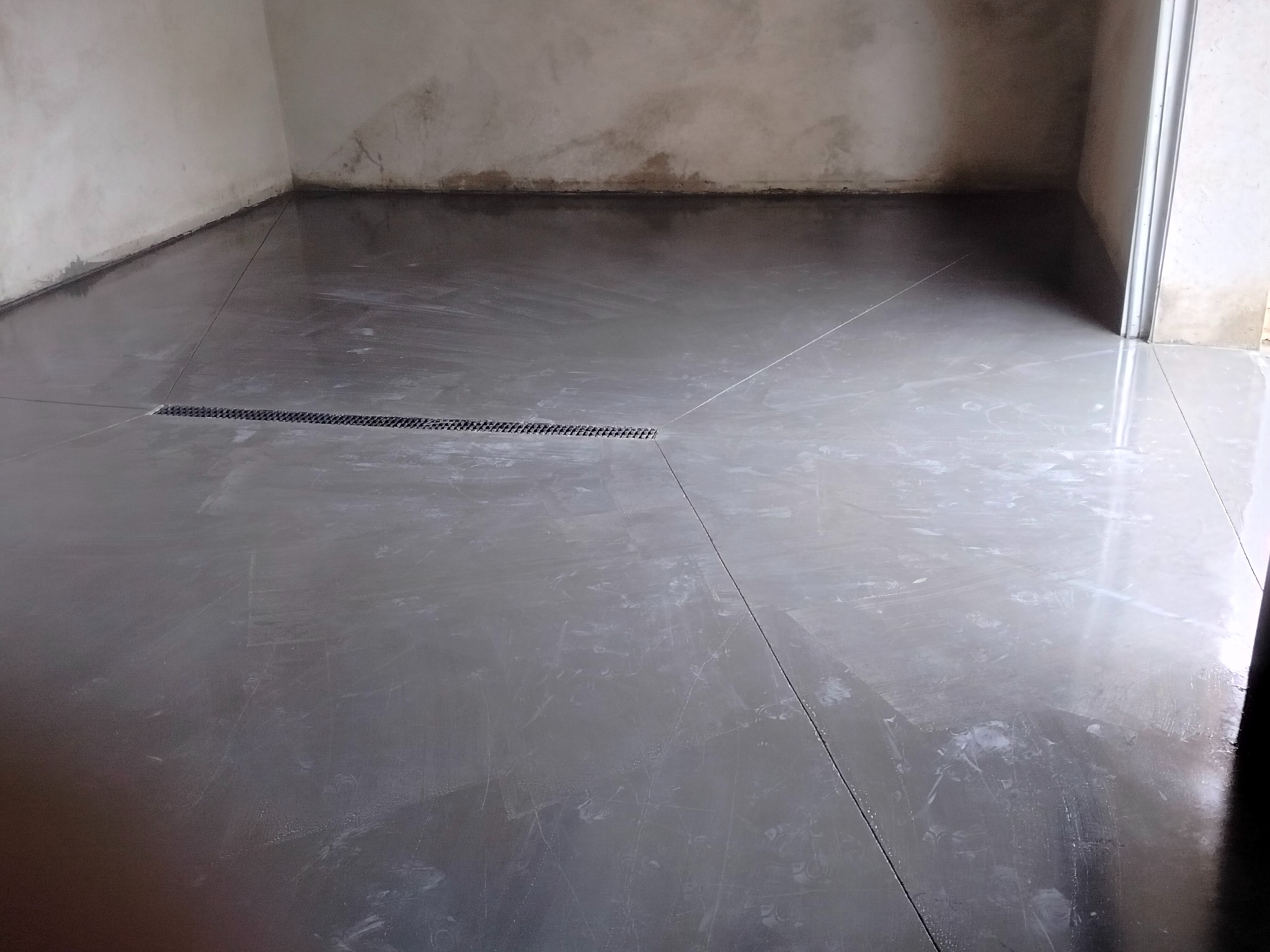 Świeżo wylana, błyszcząca posadzka przemysłowa z widocznym odpływem liniowym i śladami po narzędziach, w surowym wnętrzu.