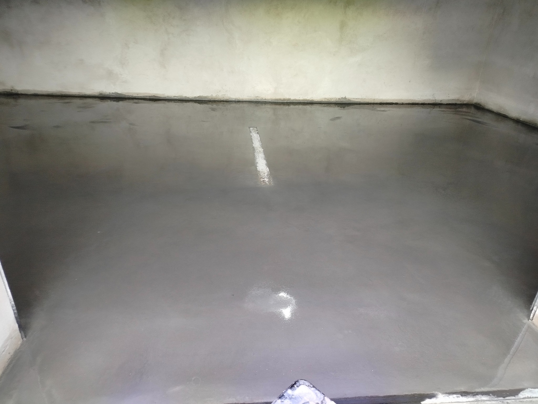 Świeżo wylana, gładka, szara posadzka w narożniku pomieszczenia, z widocznym fragmentem listwy przypodłogowej i odblaskami światła na powierzchni.