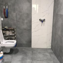 BEM HOUSE ARKADIUSZ BEMBENEK - Nowoczesna łazienka po renowacji z szarymi i białymi płytkami, widoczny podwieszany sedes z otwartą klapą i instalacją hydrauliczną, oraz pojemnik z farbą przy drzwiach.