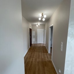 BEM HOUSE ARKADIUSZ BEMBENEK - Wykończony korytarz w mieszkaniu z drewnopodobną podłogą, jasnymi ścianami i nowoczesnym oświetleniem sufitowym. Widoczne drzwi wejściowe i wewnętrzne.