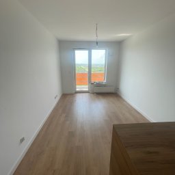 BEM HOUSE ARKADIUSZ BEMBENEK - Jasne wnętrze po remoncie: parkiet, białe ściany, okno balkonowe z widokiem na zieleń. Minimalizm i nowoczesność. Widoczne gniazdka elektryczne i wiszący kabel.