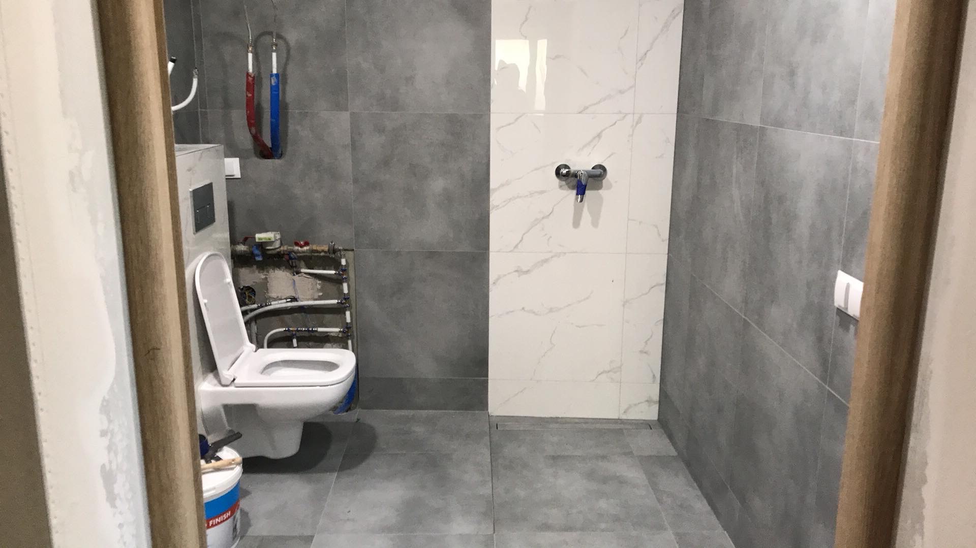 Nowoczesna łazienka po renowacji z szarymi i białymi płytkami, widoczny podwieszany sedes z otwartą klapą i instalacją hydrauliczną, oraz pojemnik z farbą przy drzwiach.