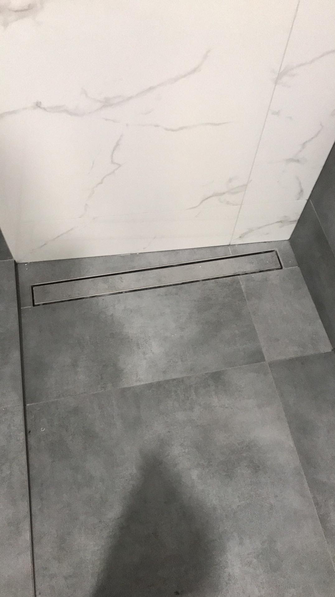 Szary gres na podłodze łazienki z liniowym odpływem pod ścianą wyłożoną białymi płytkami imitującymi marmur.