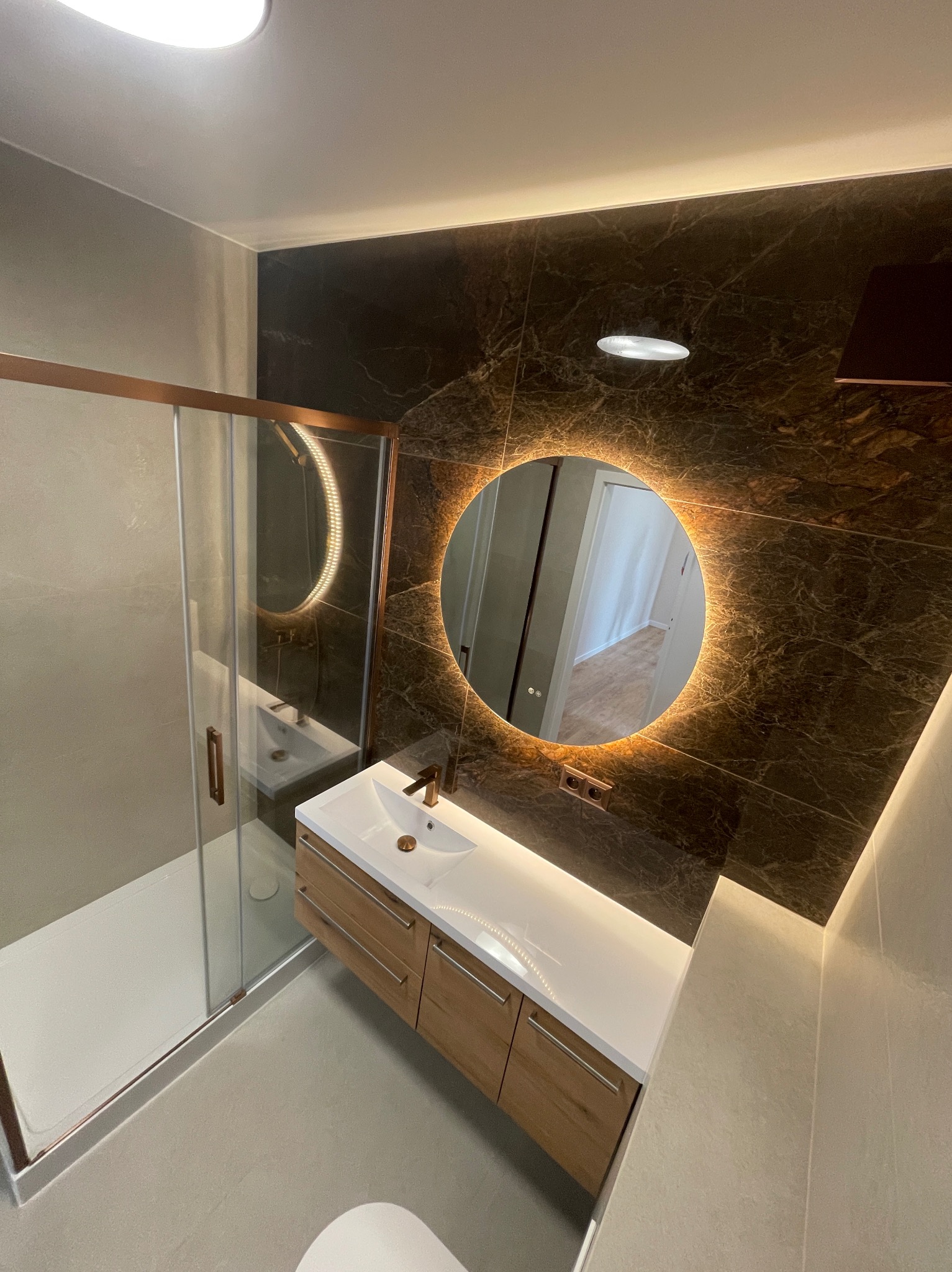 Nowoczesna łazienka z kabiną prysznicową, umywalką z szafką w drewnie i okrągłym lustrem z podświetleniem LED na tle ściany z ciemnego marmuru.