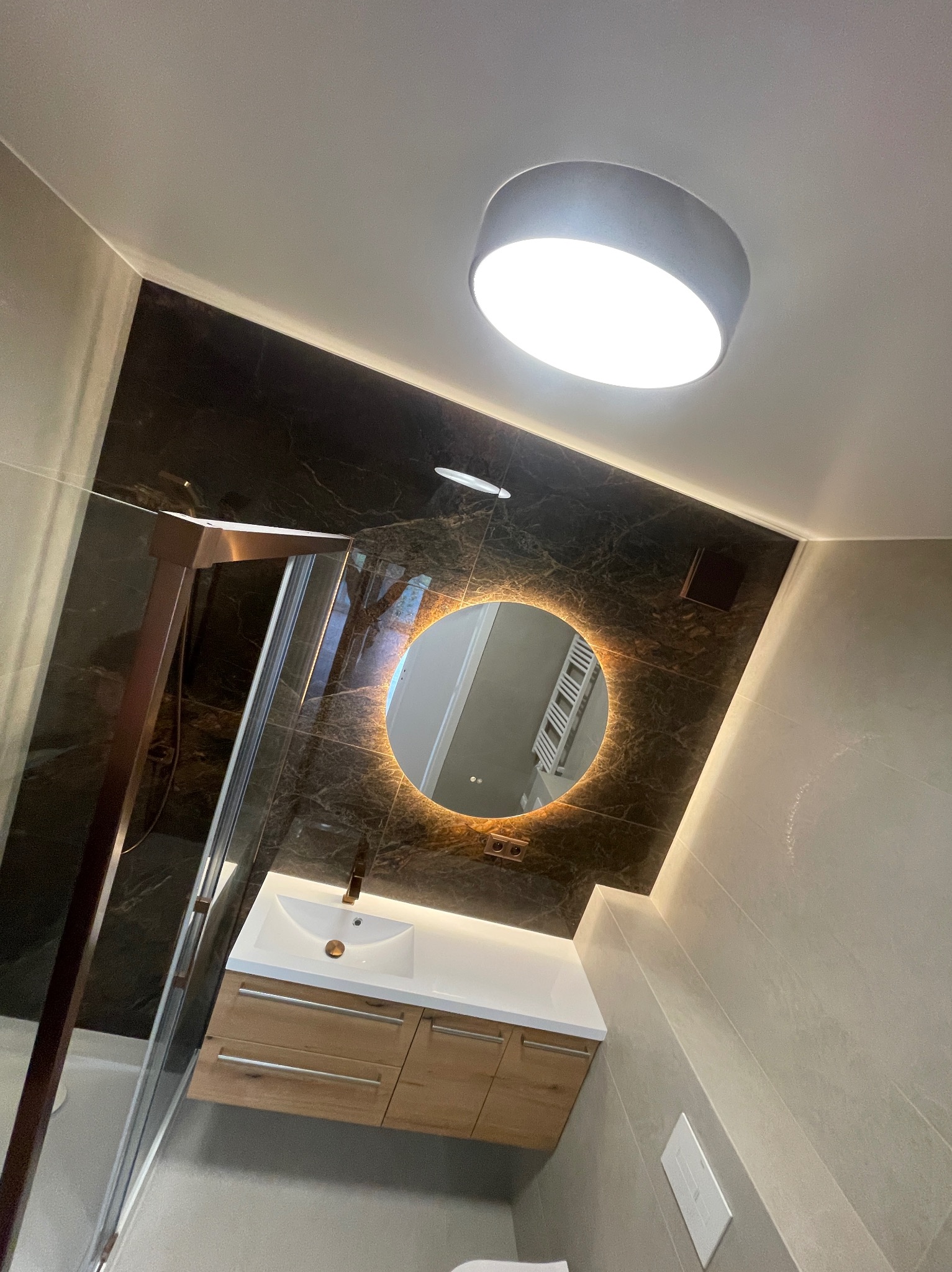 Nowoczesna łazienka z kabiną prysznicową, umywalką z szafką i okrągłym lustrem z podświetleniem LED na czarnej, marmurkowej ścianie. Oświetlenie sufitowe.