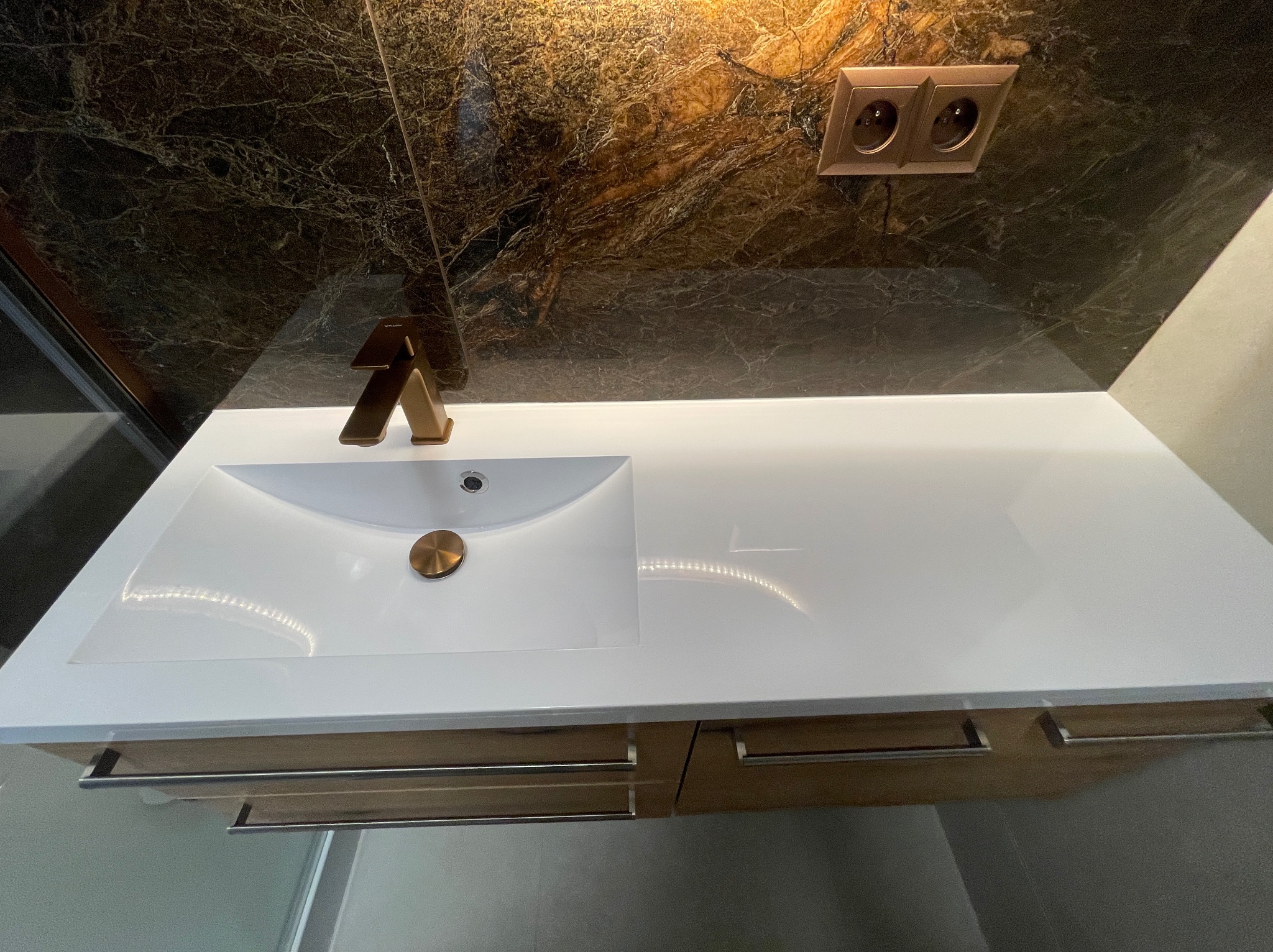 Biała umywalka z armaturą w odcieniach złota, na tle ściany z marmurowym wzorem i gniazdkami elektrycznymi. Nowoczesne wykończenie łazienki.