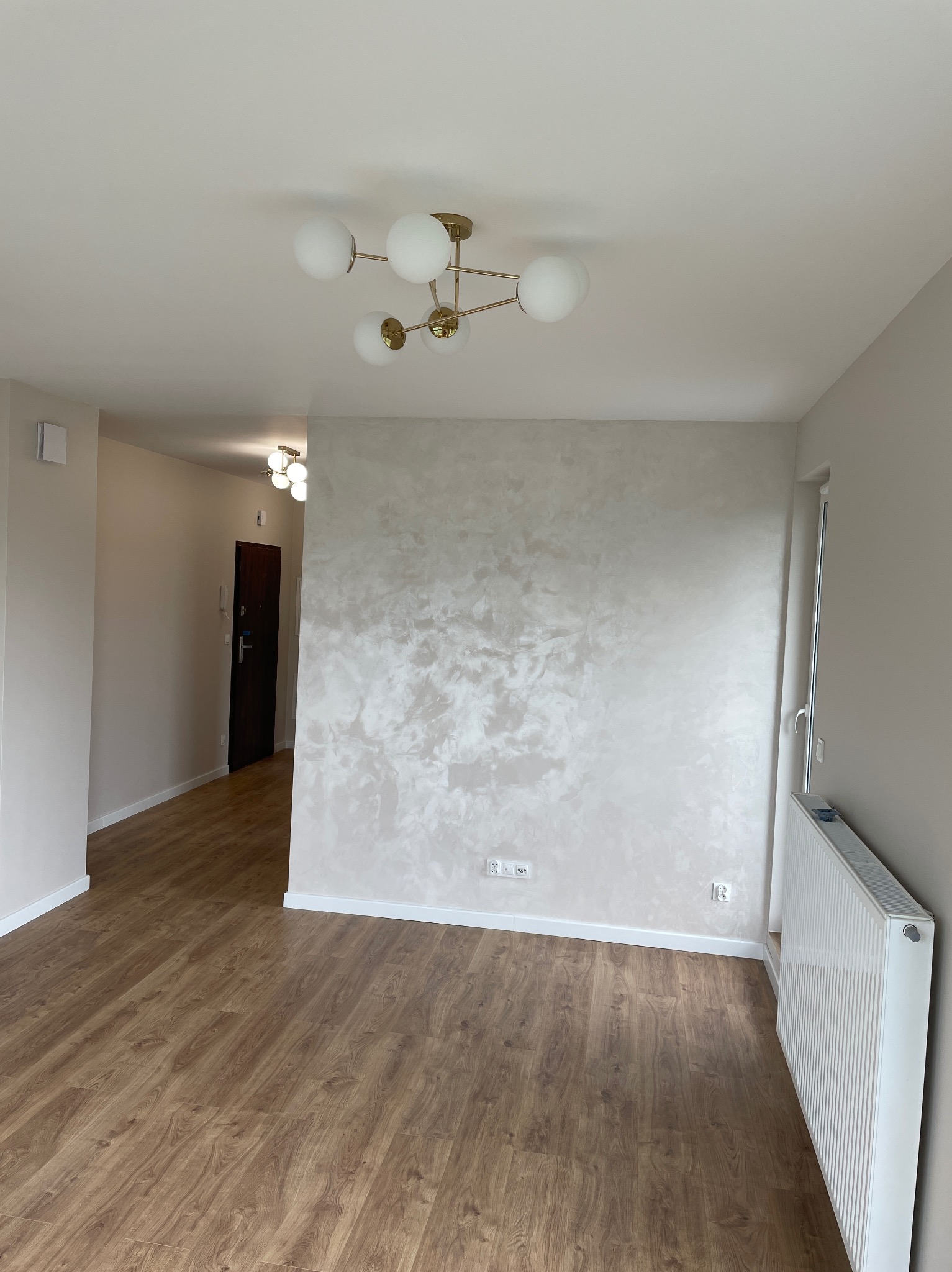 Wykończone wnętrze w Głogowie Małopolskim: drewnopodobna podłoga, ściana z efektem dekoracyjnym, nowoczesna lampa sufitowa. Widok na korytarz i grzejnik.