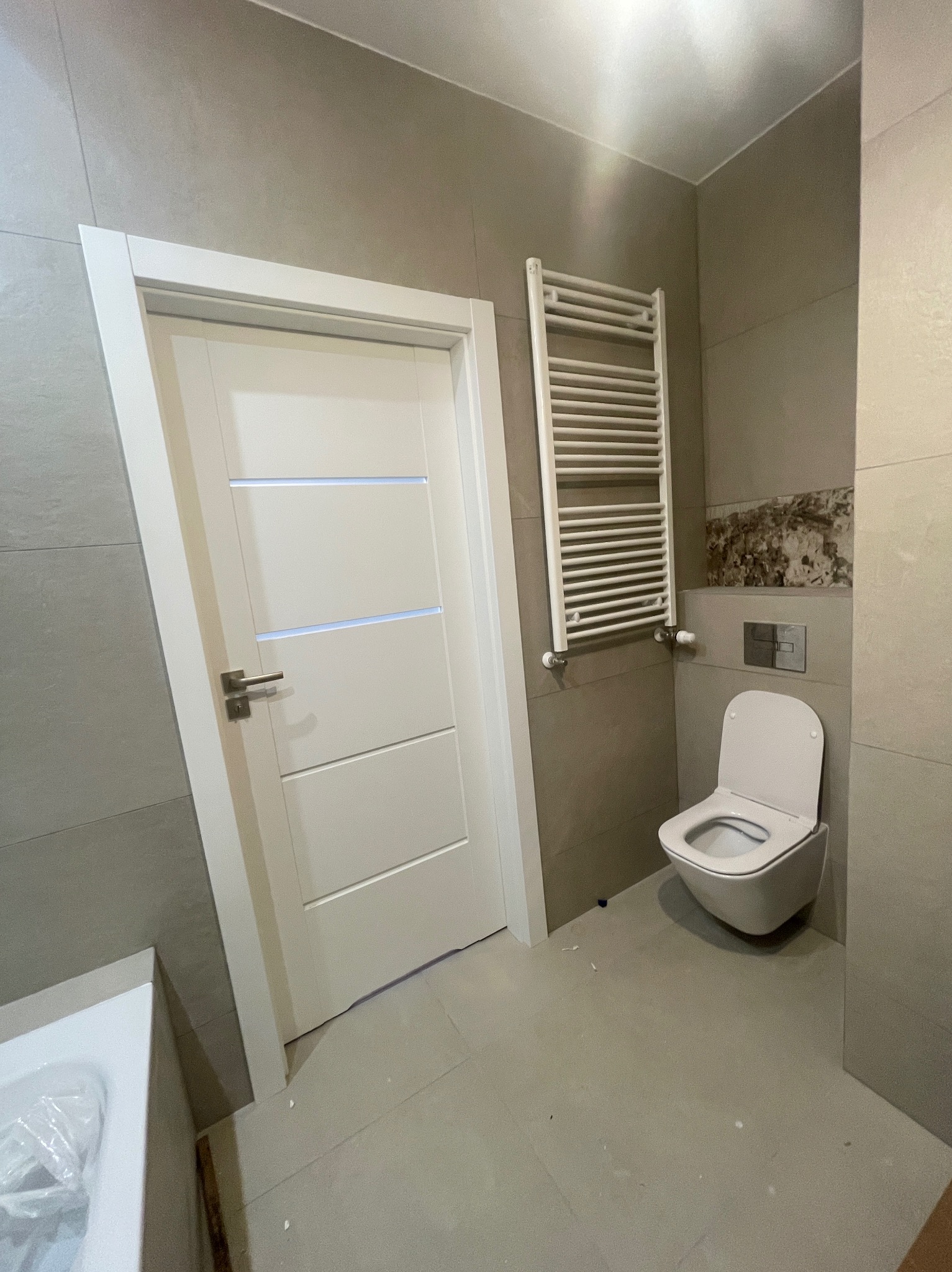 Nowoczesna łazienka z beżowymi płytkami, białą wiszącą toaletą i grzejnikiem drabinkowym. Białe drzwi z poziomymi frezami, zlew częściowo widoczny po lewej.