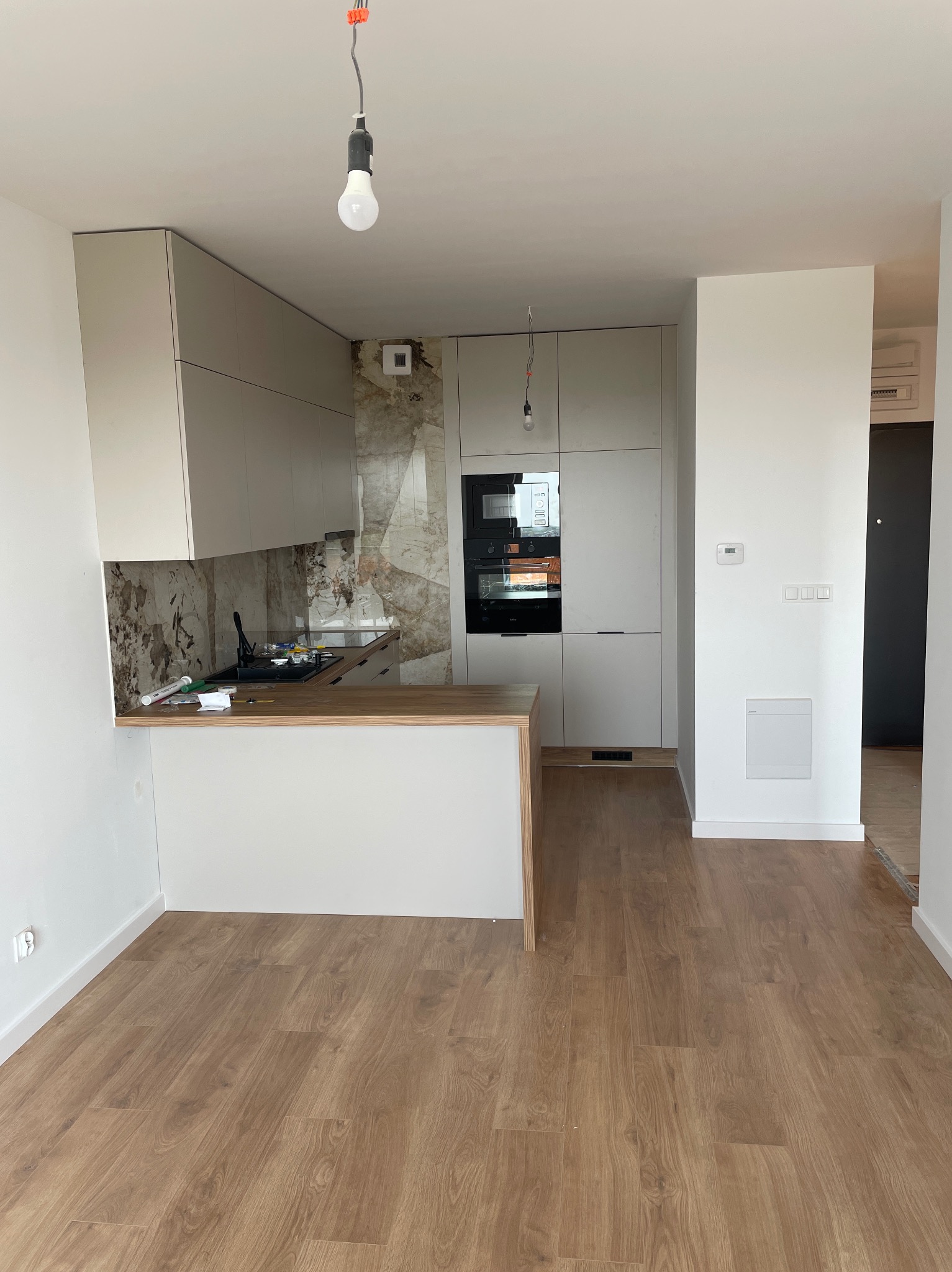 Jasna, nowoczesna kuchnia po remoncie z drewnianym blatem i marmurowym backsplashiem. Widoczne nowe panele podłogowe i niedokończone podłączenie oświetlenia.
