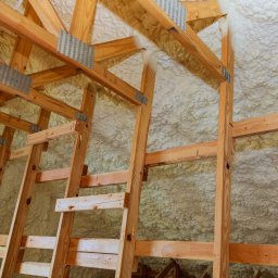 KYORA SPÓŁKA Z OGRANICZONĄ ODPOWIEDZIALNOŚCIĄ - Poddasze z drewnianą konstrukcją krokwiową pokryte grubą warstwą jasnej pianki PUR z widocznymi metalowymi łącznikami.