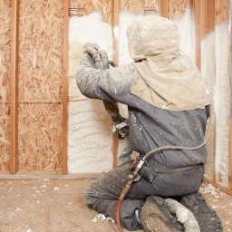 KYORA SPÓŁKA Z OGRANICZONĄ ODPOWIEDZIALNOŚCIĄ - Pracownik w kombinezonie ochronnym aplikuje piankę izolacyjną na ścianę szkieletową wewnątrz budynku, widoczne drewniane elementy konstrukcyjne, fragment instalacji elektrycznej i resztki pianki...