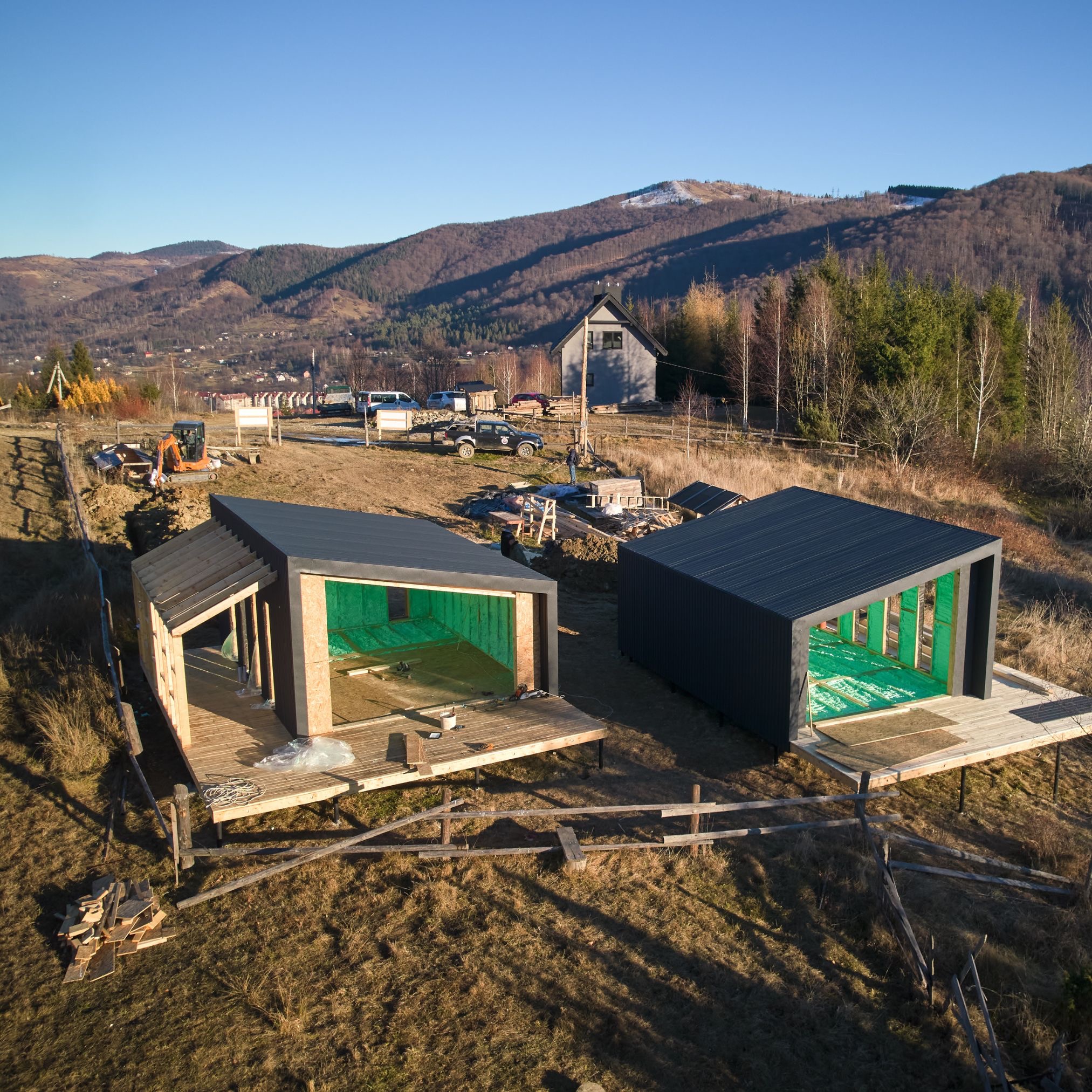Dwa szkieletowe domki modułowe na drewnianych platformach z widoczną zieloną izolacją, w tle górzysty krajobraz i zabudowa, na pierwszym planie drewniane ogrodzenie i teren budowy.