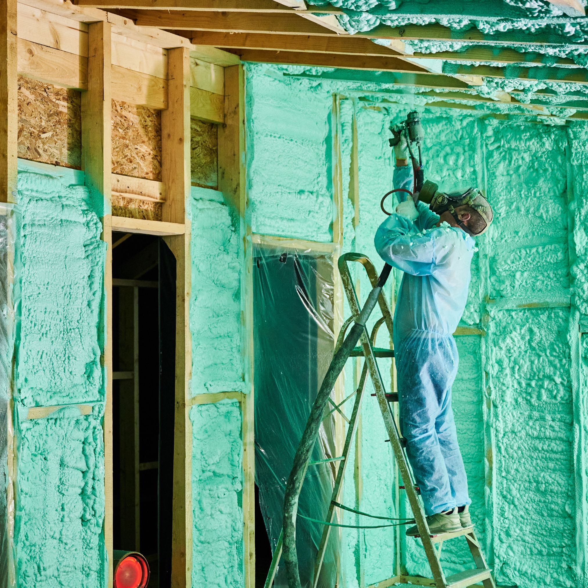 Pracownik w kombinezonie ochronnym aplikuje piankę izolacyjną na ściany szkieletowe budynku, stojąc na aluminiowej drabinie; widoczny fragment niezabezpieczonego otworu okiennego.