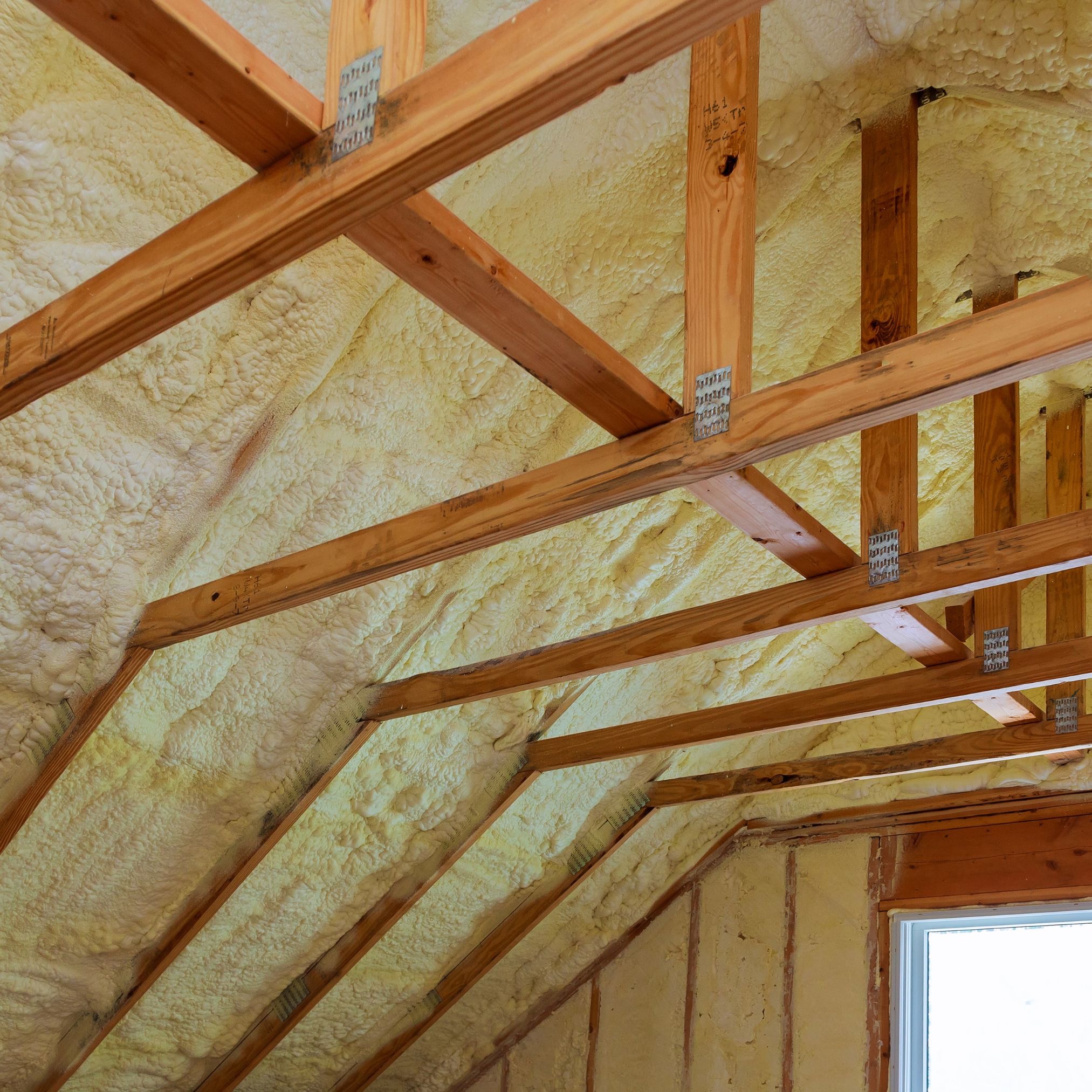 Poddasze z widoczną konstrukcją drewnianą pokryte żółtawą pianą izolacyjną, mocowaną do krokwi za pomocą metalowych łączników.