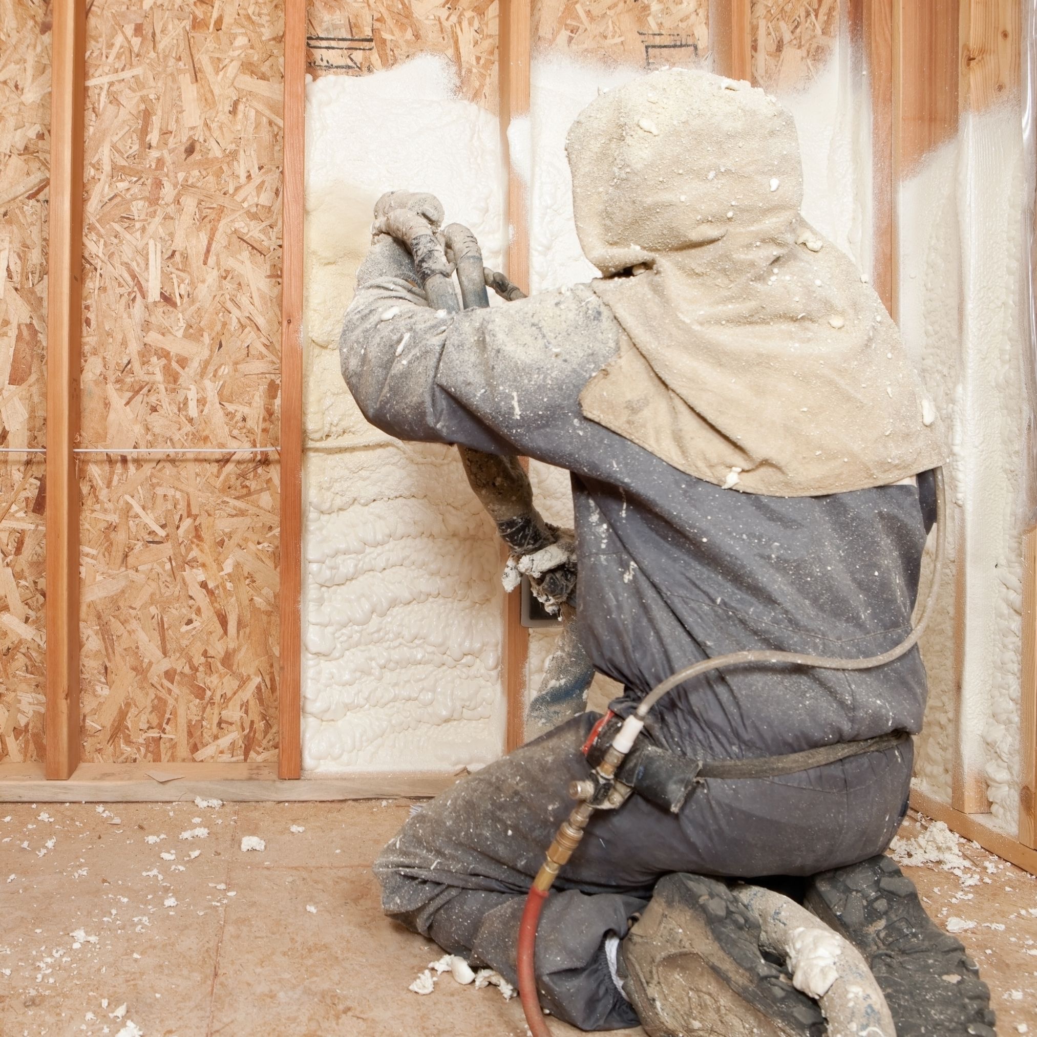 Pracownik w kombinezonie ochronnym aplikuje piankę izolacyjną na ścianę szkieletową wewnątrz budynku, widoczne drewniane elementy konstrukcyjne, fragment instalacji elektrycznej i resztki pianki...