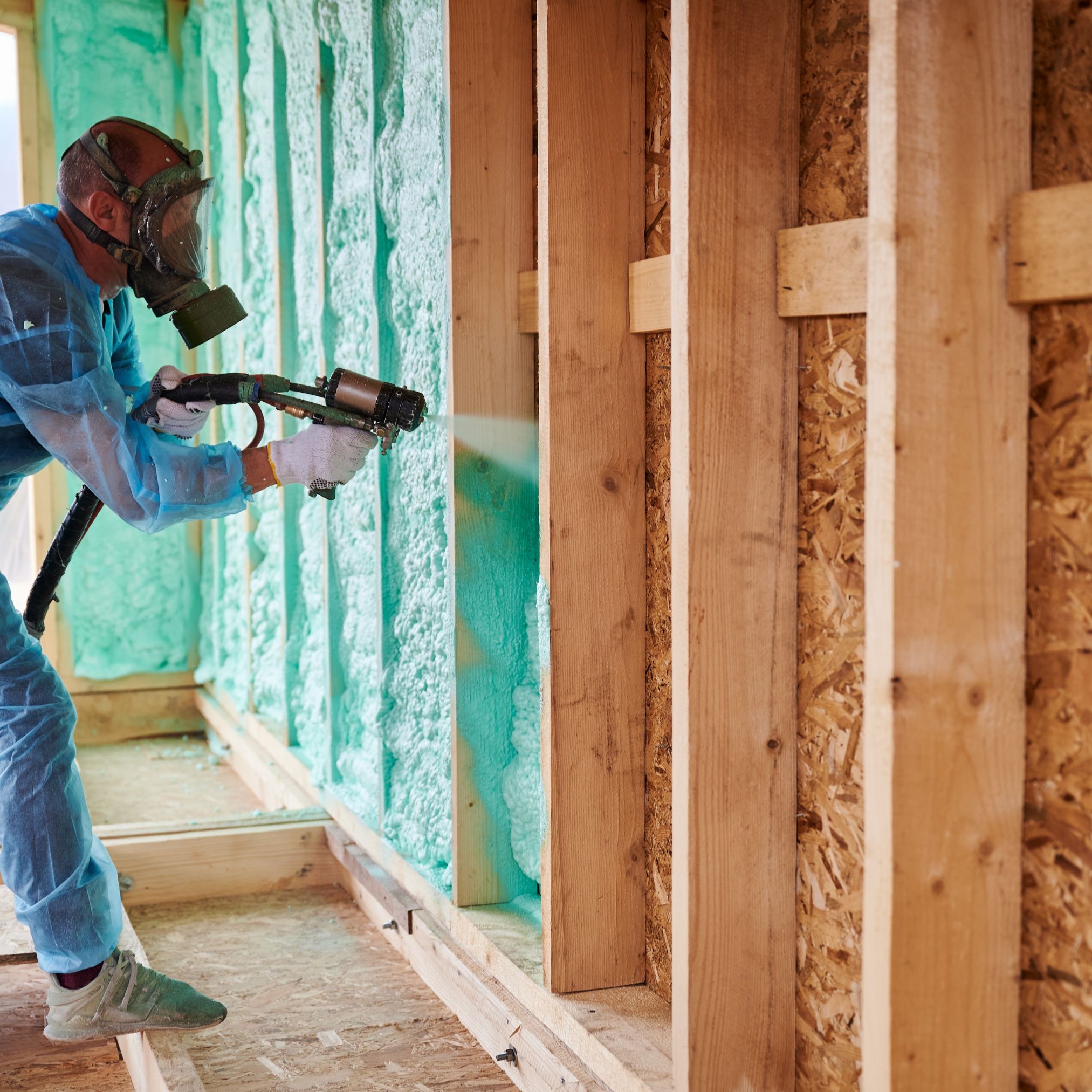 Pracownik w kombinezonie i masce gazowej aplikuje pianę izolacyjną na ściany budynku w trakcie budowy, widoczne drewniane elementy konstrukcyjne.