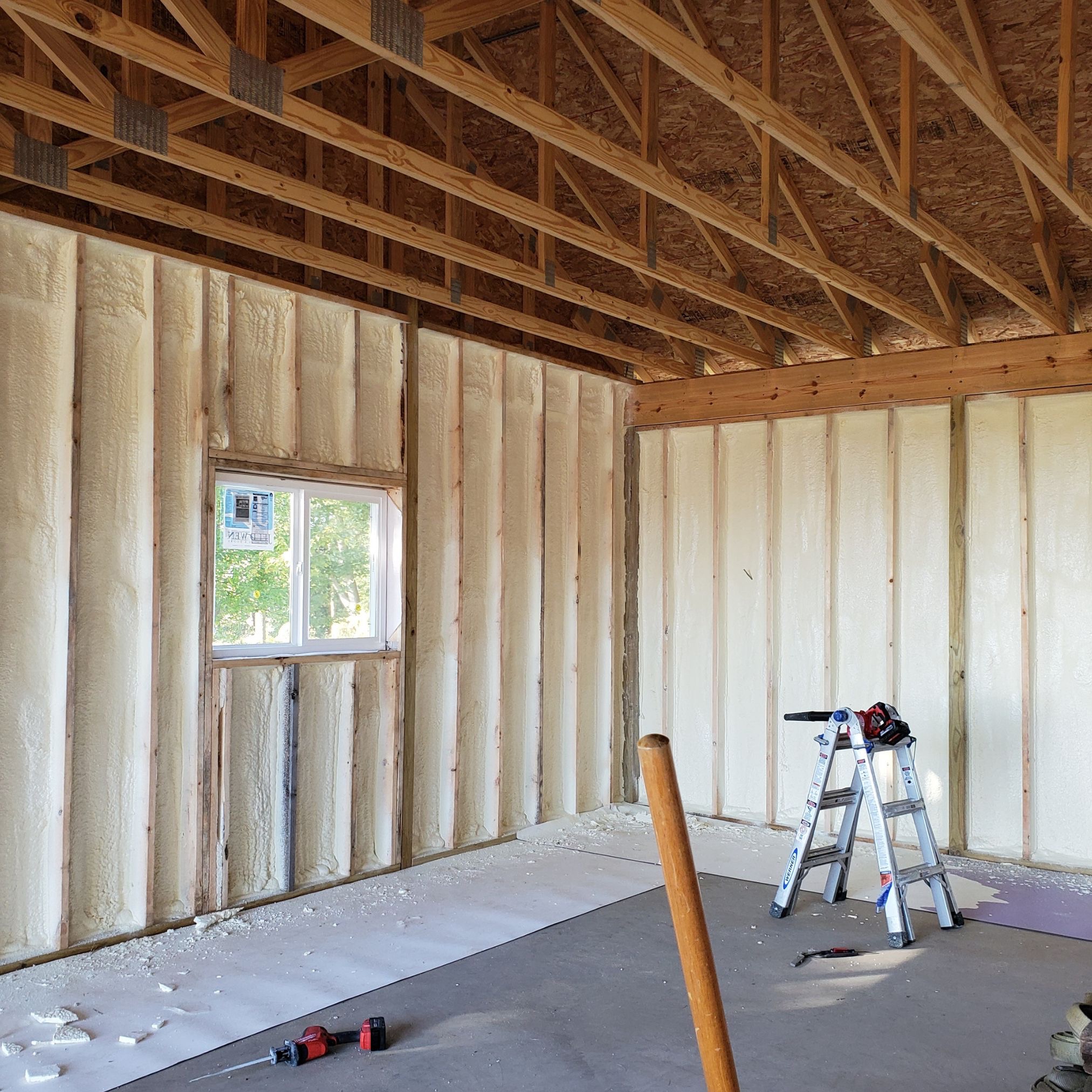Wnętrze budynku w trakcie ocieplania: ściany wypełnione pianką izolacyjną, widoczne drewniane belki konstrukcyjne, okno z widokiem na zieleń, drabina aluminiowa z narzędziami oraz resztki pianki...