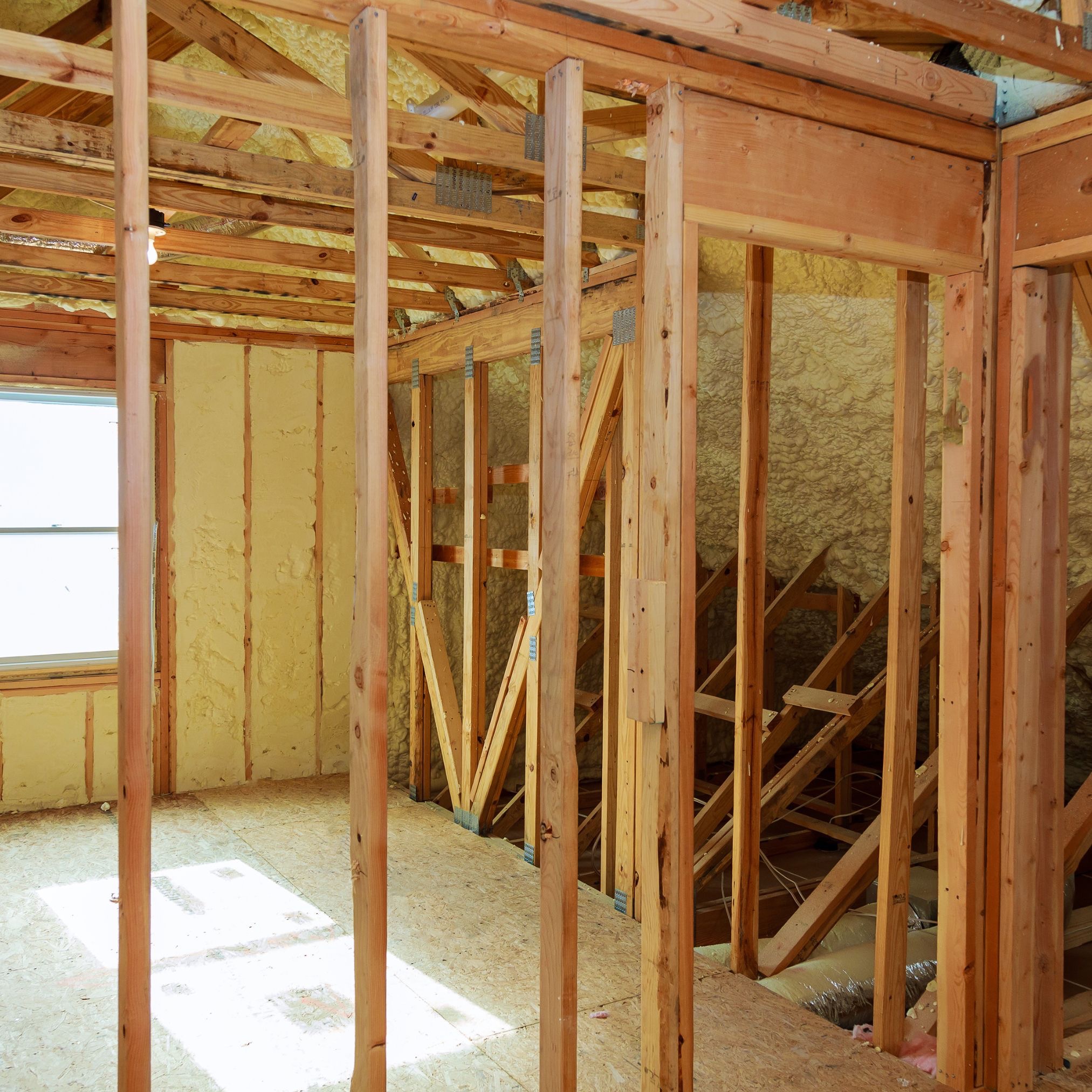 Poddasze w trakcie izolacji: widoczne drewniane elementy konstrukcyjne, ściany i dach pokryte żółtą pianą izolacyjną, okno w tle wpuszcza naturalne światło.
