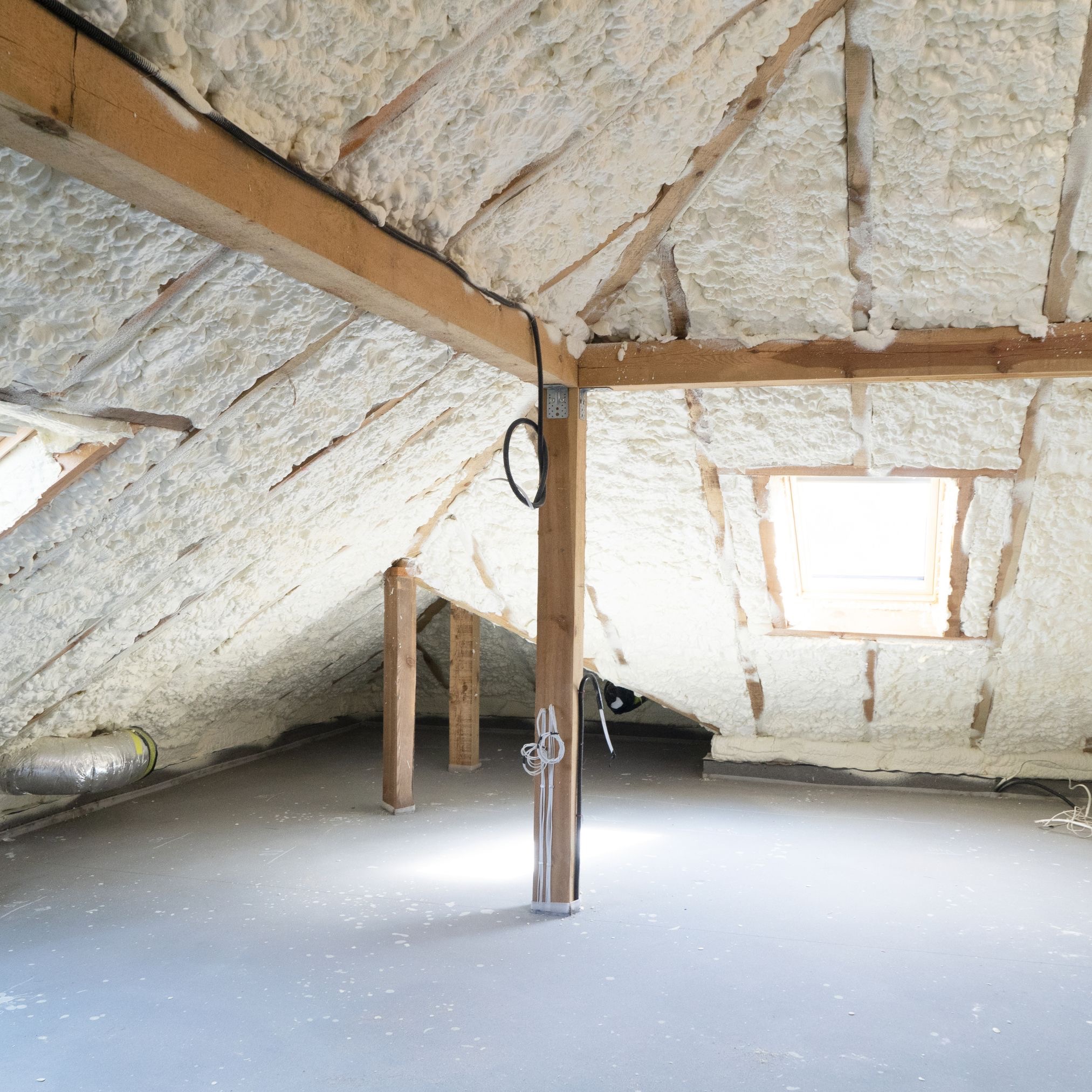 Poddasze z drewnianą konstrukcją pokryte grubą warstwą białej piany izolacyjnej, widoczne okno dachowe i fragment instalacji elektrycznej.