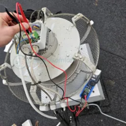 Pomiar elektryczny wentylatora rekuperatora na dachu, widoczne przewody, miernik cęgowy i elementy instalacji w szarej obudowie z metalową siatką ochronną.