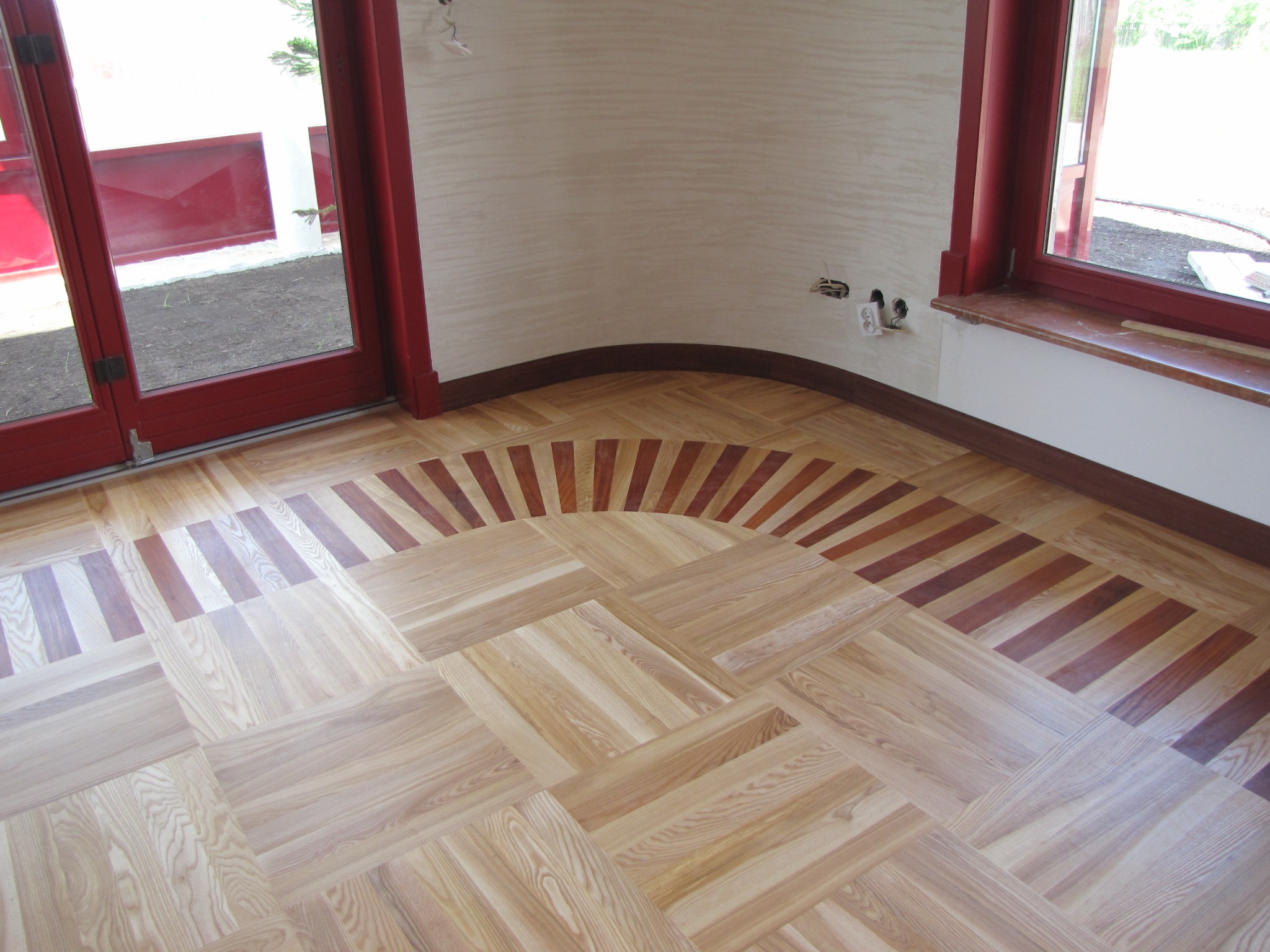 Ułożony parkiet w jodełkę z dekoracyjnym obramowaniem z ciemniejszych desek, widoczne czerwone ramy okienne i drzwi balkonowych, ściana z delikatną fakturą.