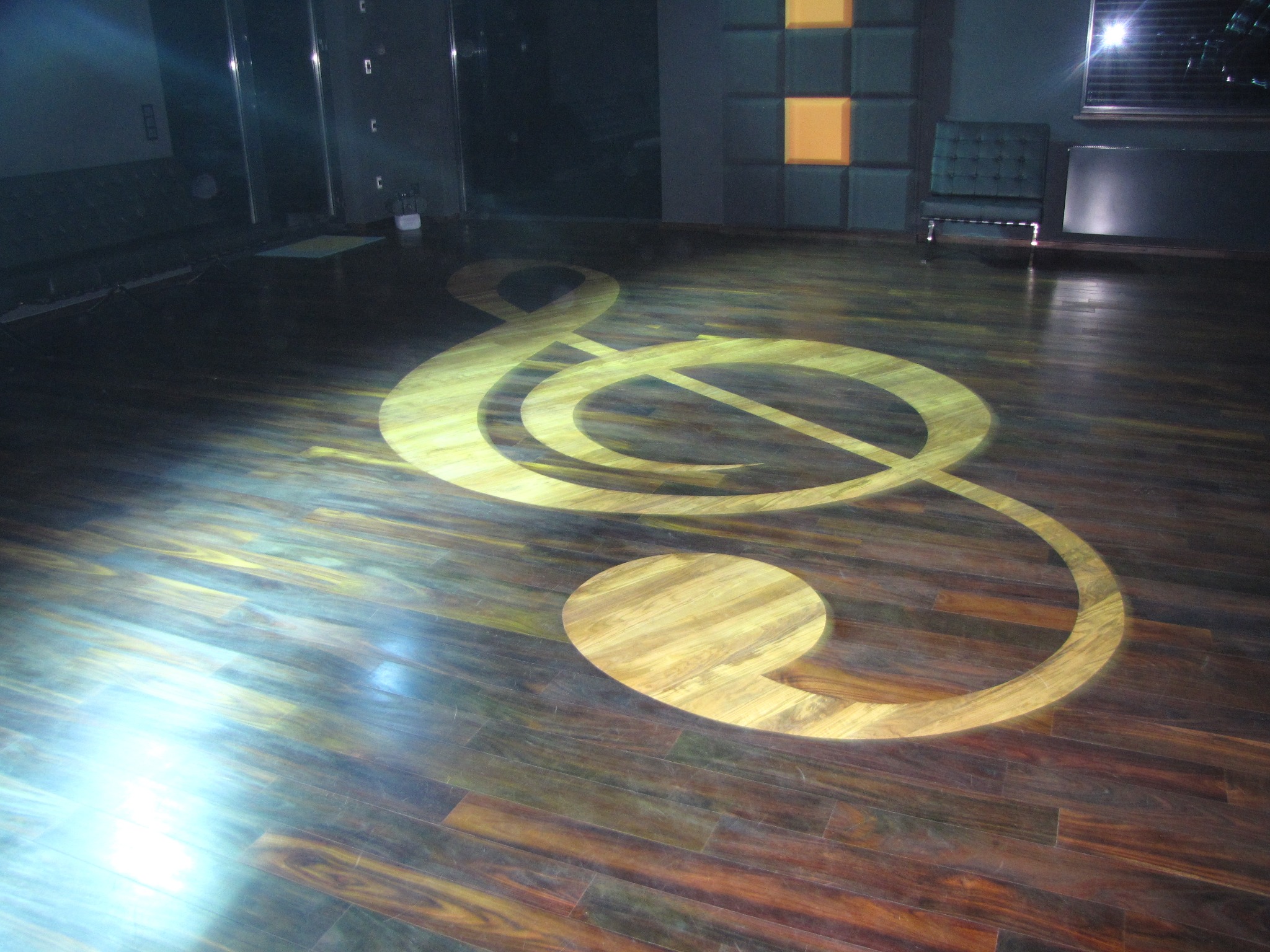 Parkiet z ciemnego drewna z intarsją w kształcie klucza wiolinowego, oświetlony punktowo, w tle minimalistyczne wnętrze z ciemnymi ścianami i designerskim fotelem.