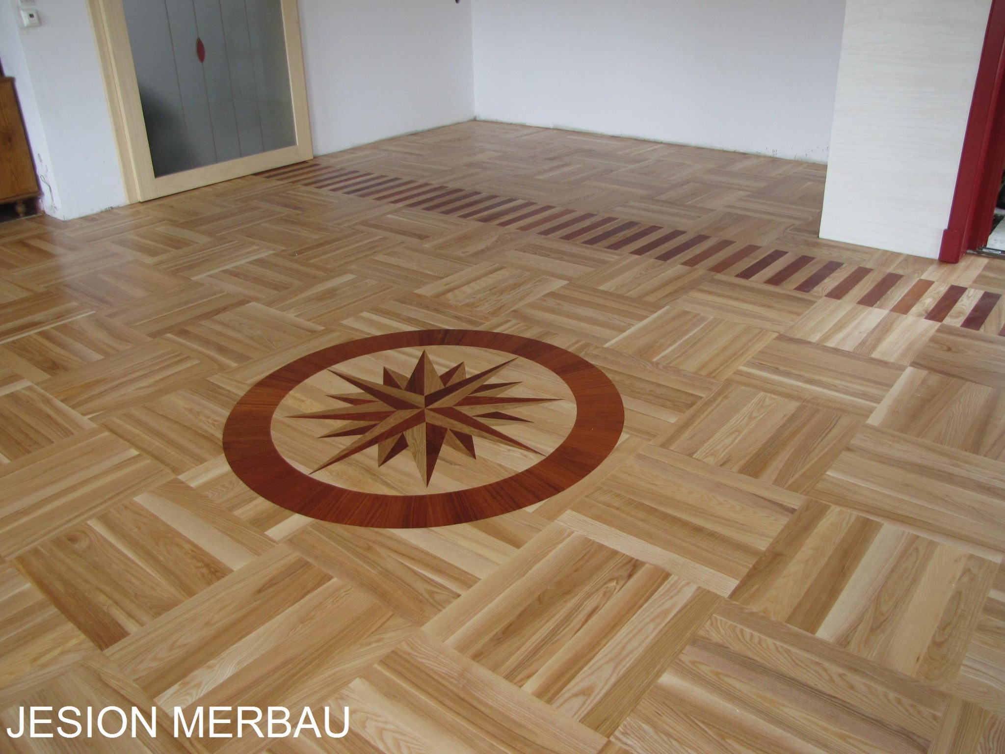 Parkiet z jasnego drewna ułożony we wzór kwadratów, z dekoracyjnym motywem rozety w centrum i bordiurą z ciemniejszego drewna przy ścianie. Widoczny fragment drzwi z szybą w tle.