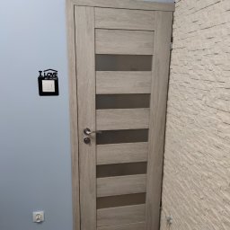 ROB-BUD - Zamontowane drzwi wewnętrzne z przeszkleniami, w odcieniach szarości, na tle niebieskiej ściany z dekoracją 'I love my home' nad włącznikiem światła i ściany wyłożonej dekoracyjną białą cegłą.