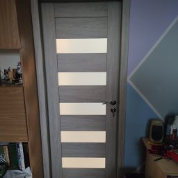 ROB-BUD - Zamknięte, szare drzwi wewnętrzne z poziomymi przeszkleniami, widoczne na tle ściany w geometryczne wzory i fragmentu drewnianego regału.