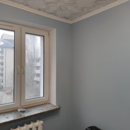 ROB-BUD - Pomalowane na szaro ściany w pokoju z ozdobnym sufitem, białe okno z widokiem na blok i grzejnik pod parapetem. Na podłodze czarna folia ochronna.