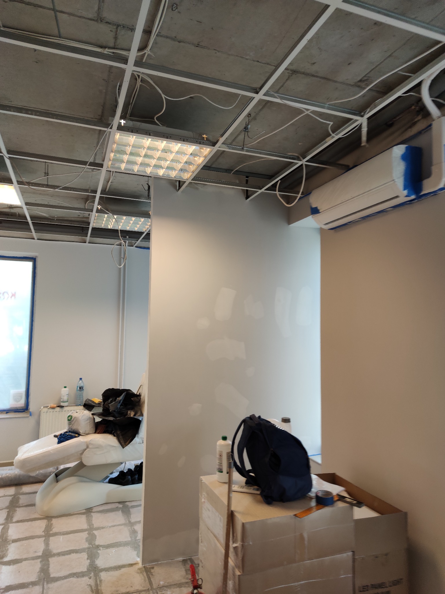 Wnętrze biura w trakcie prac wykończeniowych: surowy sufit z widoczną instalacją, świeżo pomalowana ściana, klimatyzator zabezpieczony taśmą malarską, podłoga przygotowana do położenia paneli...