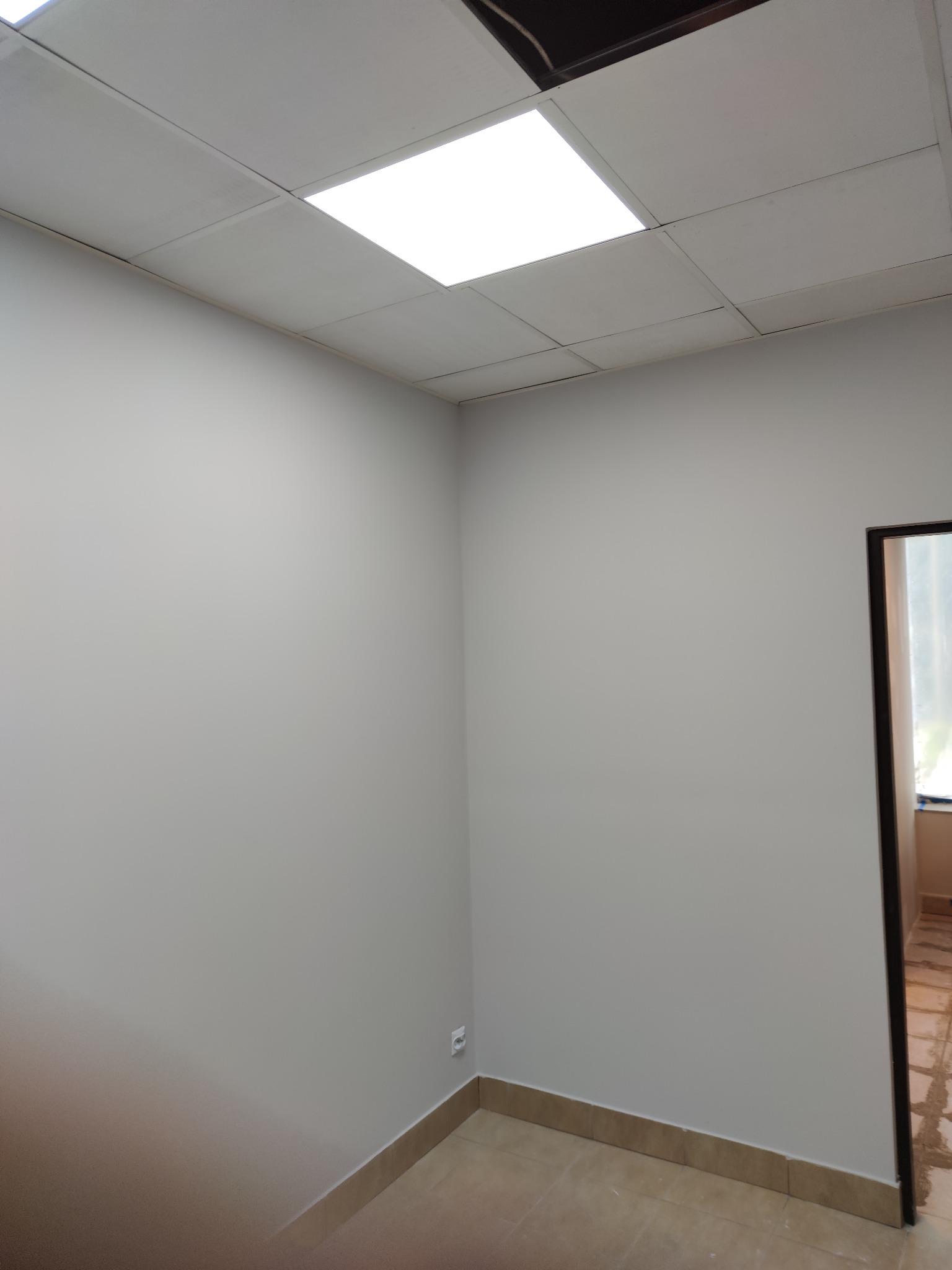 Szare, świeżo pomalowane ściany w rogu pokoju biurowego z widoczną instalacją oświetleniową w suficie podwieszanym i fragmentem futryny drzwiowej. Widoczne listwy przypodłogowe.