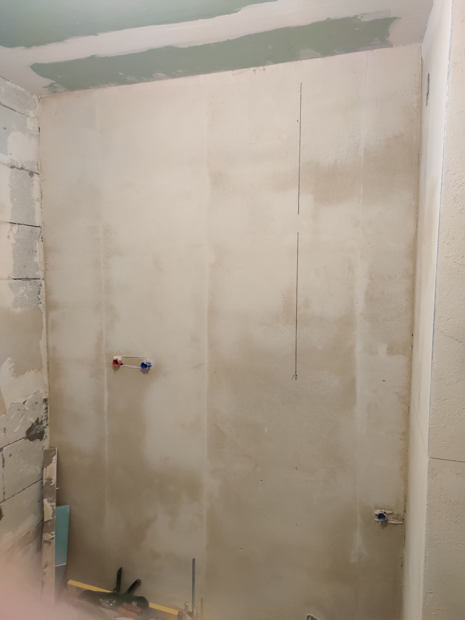 Ściana łazienki w trakcie prac przygotowawczych pod wykończenie, widoczne ślady po szpachlowaniu, pionowa linia wyznaczająca punkt odniesienia, wystające rury instalacji wodnej z zaślepkami...