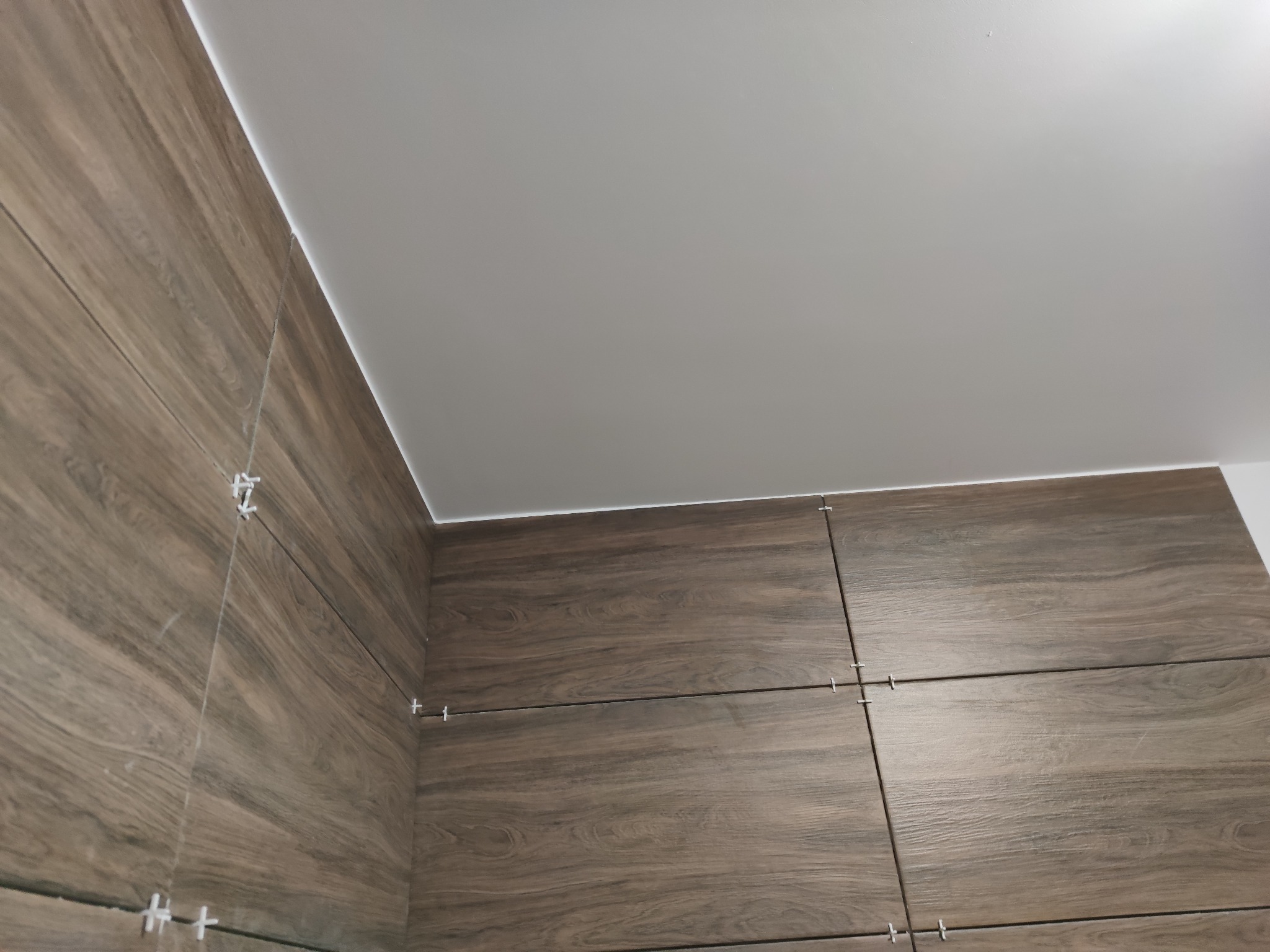 Montaż płytek imitujących drewno na ścianie w Warszawie, widoczne krzyżyki dystansowe i fragment sufitu.