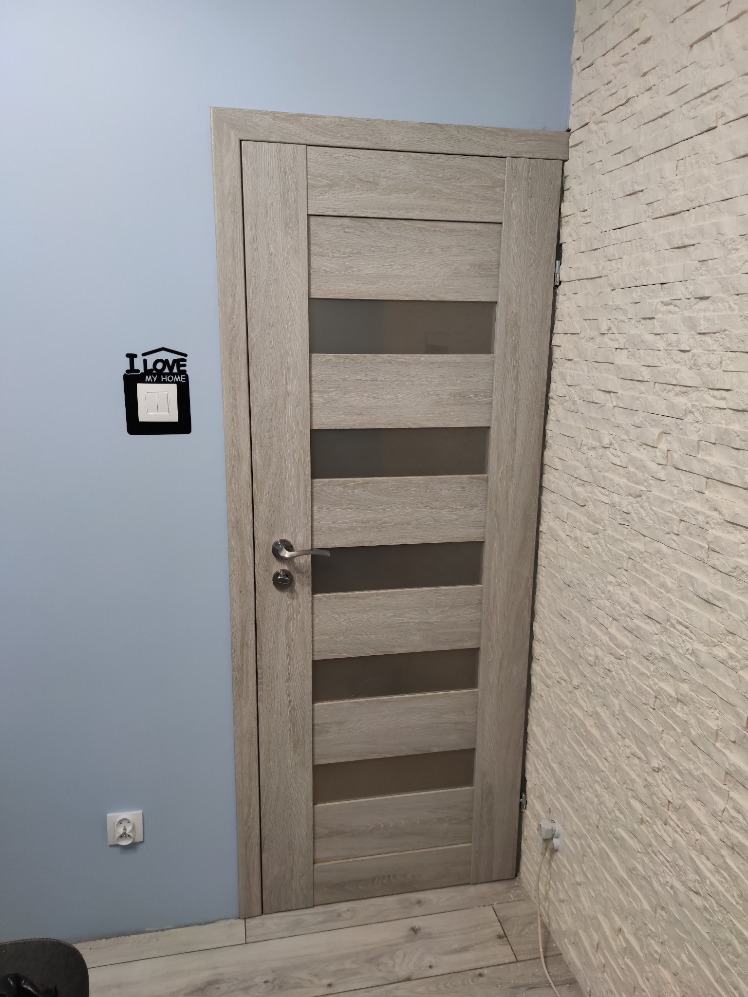 Zamontowane drzwi wewnętrzne z przeszkleniami, w odcieniach szarości, na tle niebieskiej ściany z dekoracją 'I love my home' nad włącznikiem światła i ściany wyłożonej dekoracyjną białą cegłą.