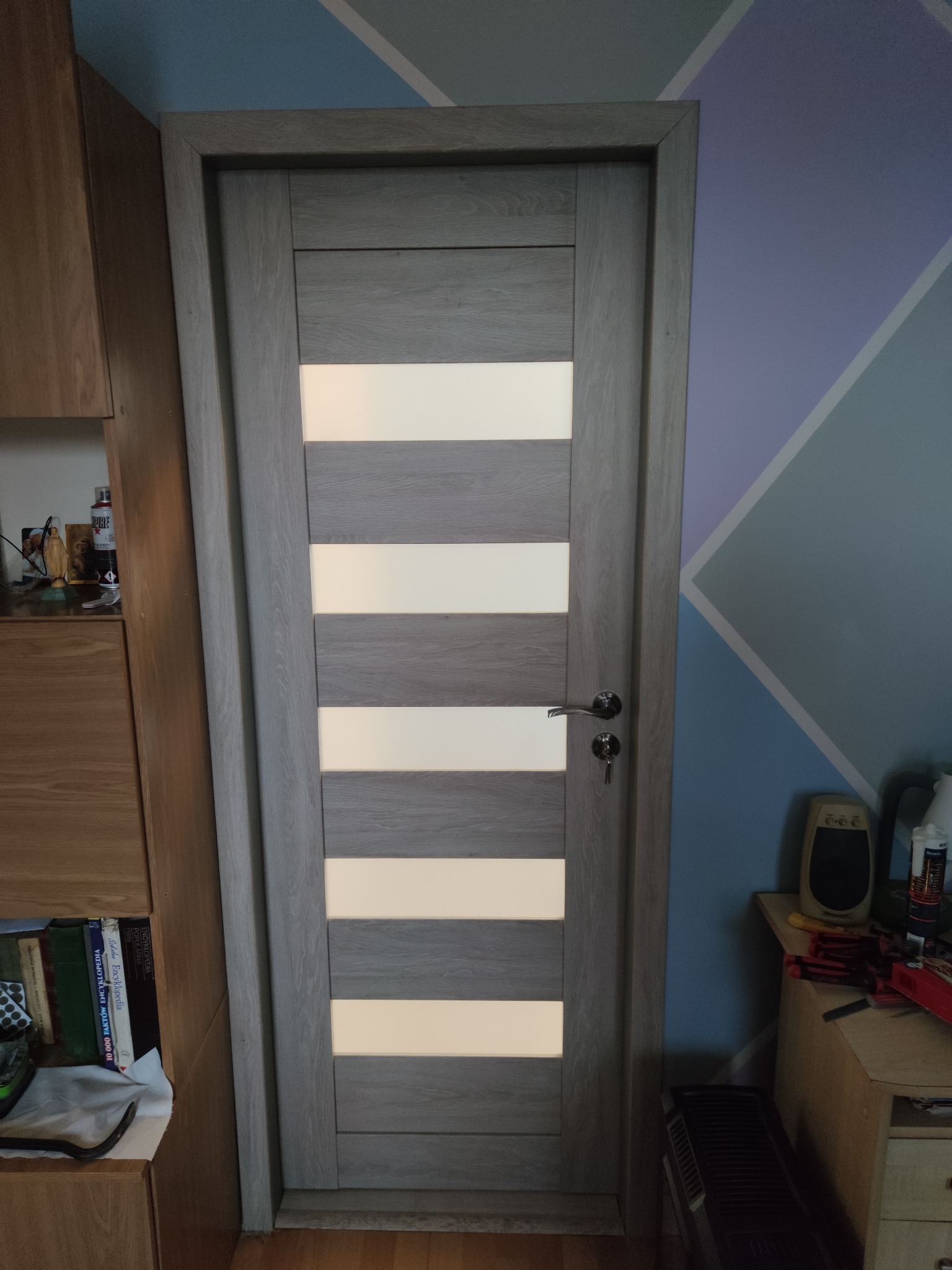 Zamknięte, szare drzwi wewnętrzne z poziomymi przeszkleniami, widoczne na tle ściany w geometryczne wzory i fragmentu drewnianego regału.
