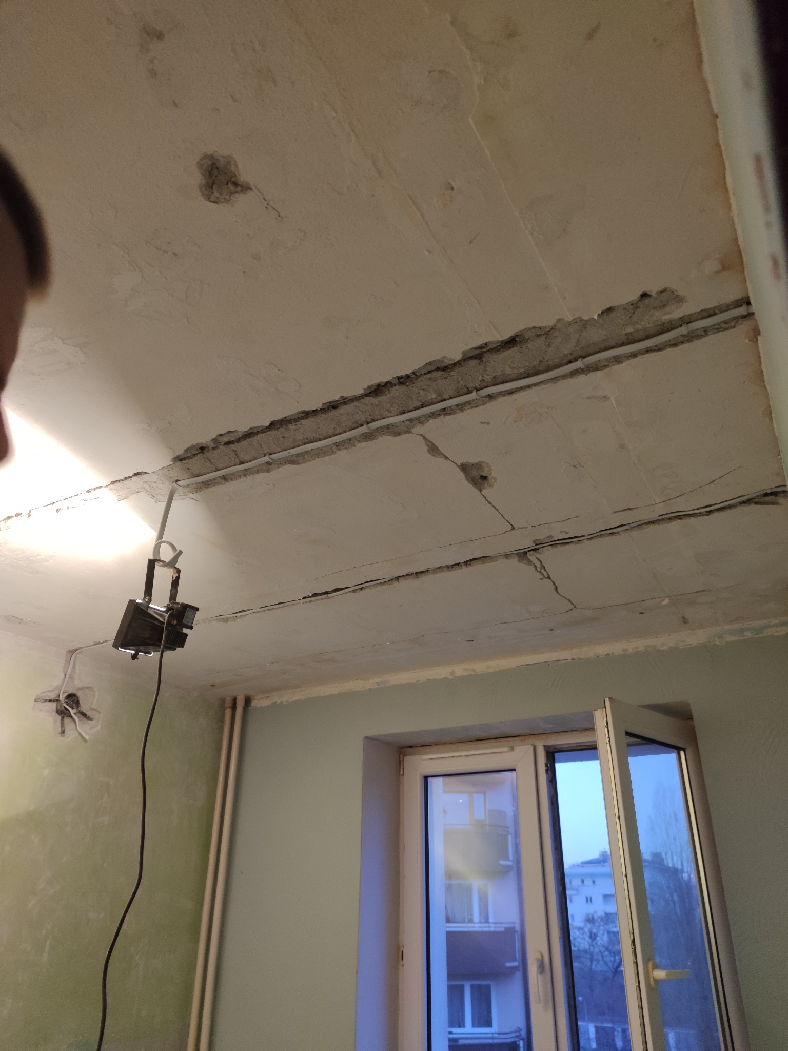 Przygotowanie sufitu do prac wykończeniowych: widoczne pęknięcia, odsłonięte przewody i lampa halogenowa oświetlająca obszar roboczy, okno w tle.