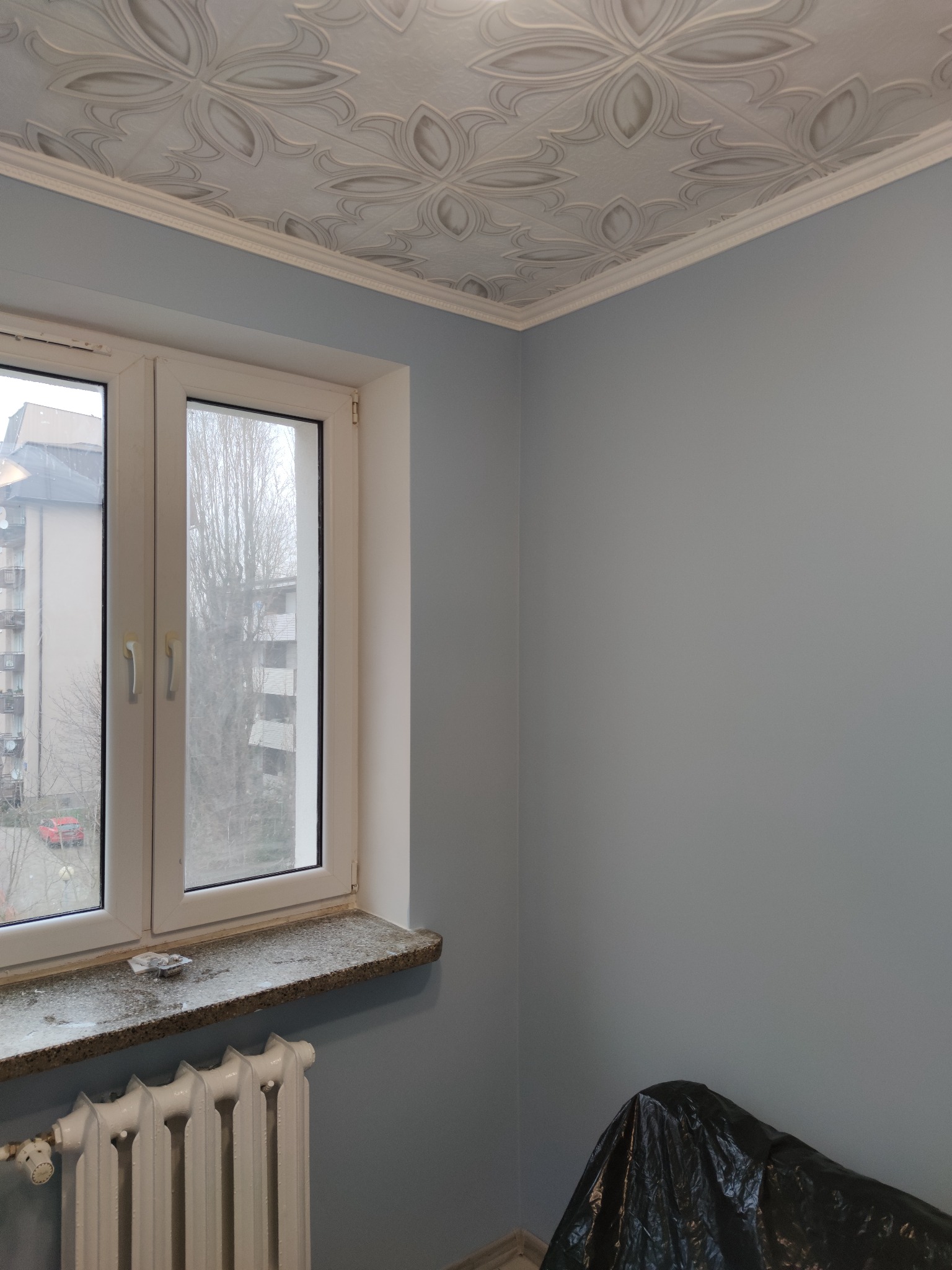Pomalowane na szaro ściany w pokoju z ozdobnym sufitem, białe okno z widokiem na blok i grzejnik pod parapetem. Na podłodze czarna folia ochronna.