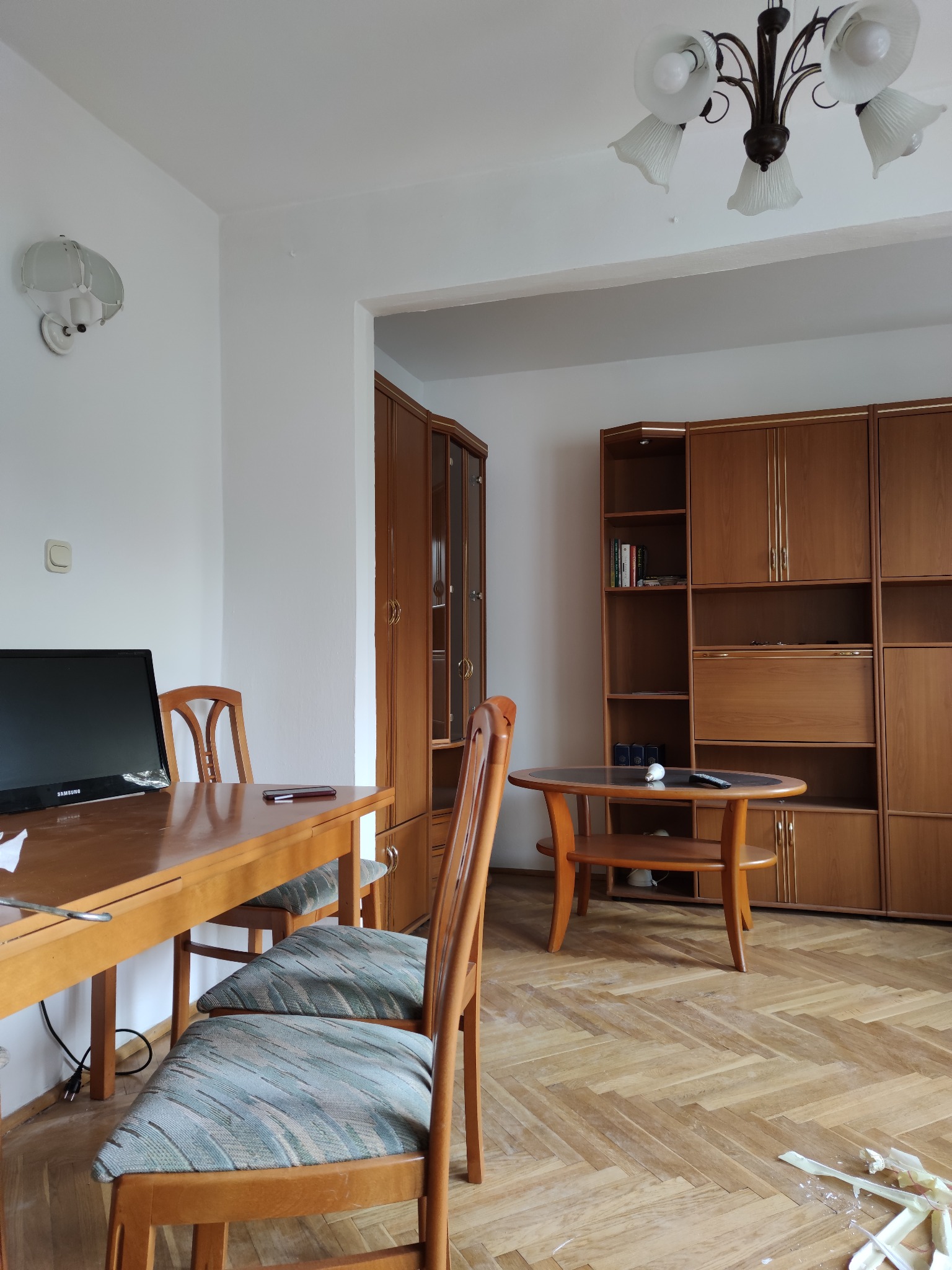 Pokój z drewnianą podłogą w jodełkę, meblami w stylu retro, lampą sufitową z kloszami, stołem z krzesłami i telewizorem; widoczne ślady prac remontowych na podłodze.