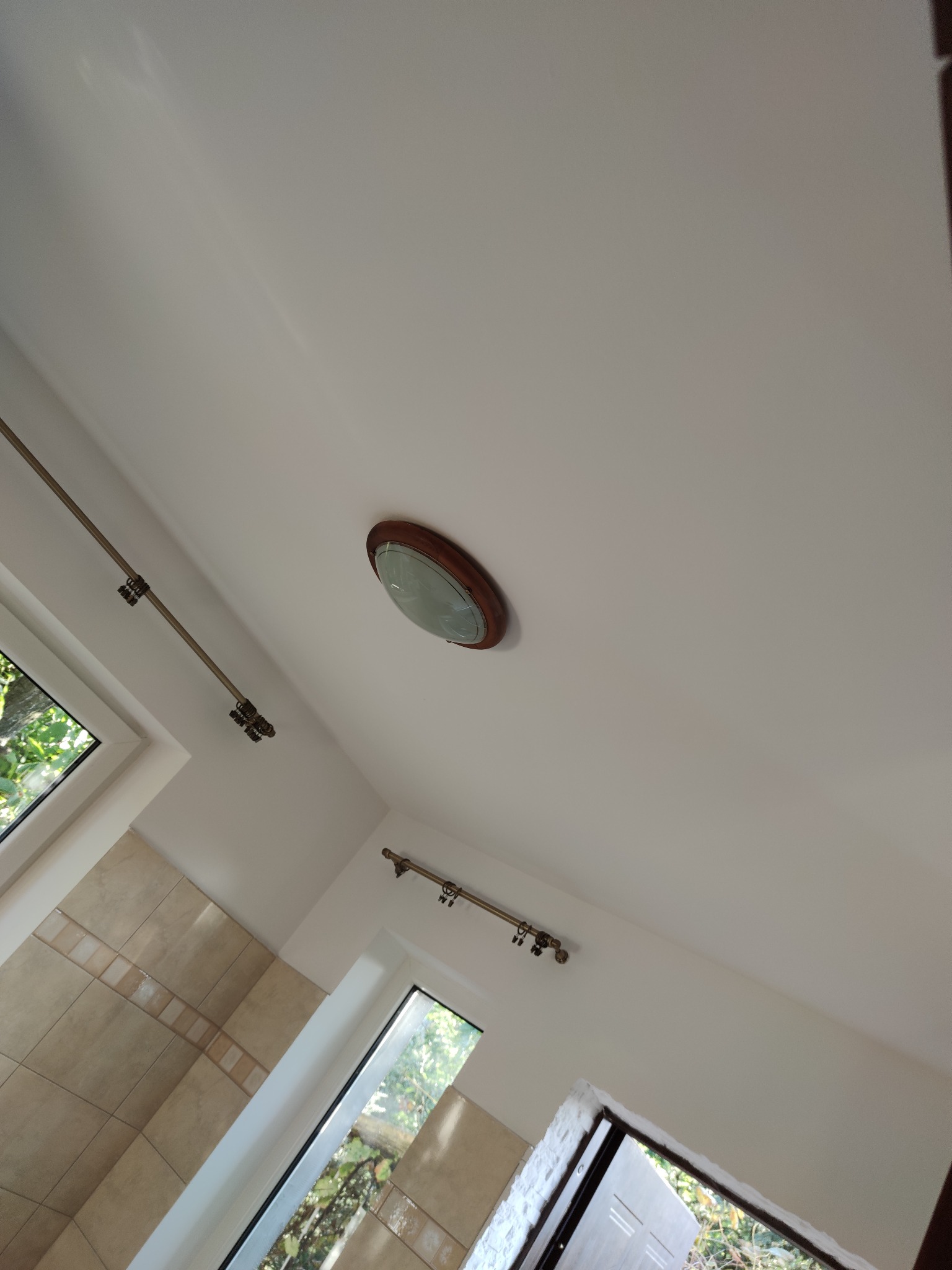 Wnętrze po odświeżeniu: biała ściana i sufit, okno z widokiem na zieleń, fragment ściany wyłożonej beżowymi płytkami i karnisz z ozdobnymi uchwytami.