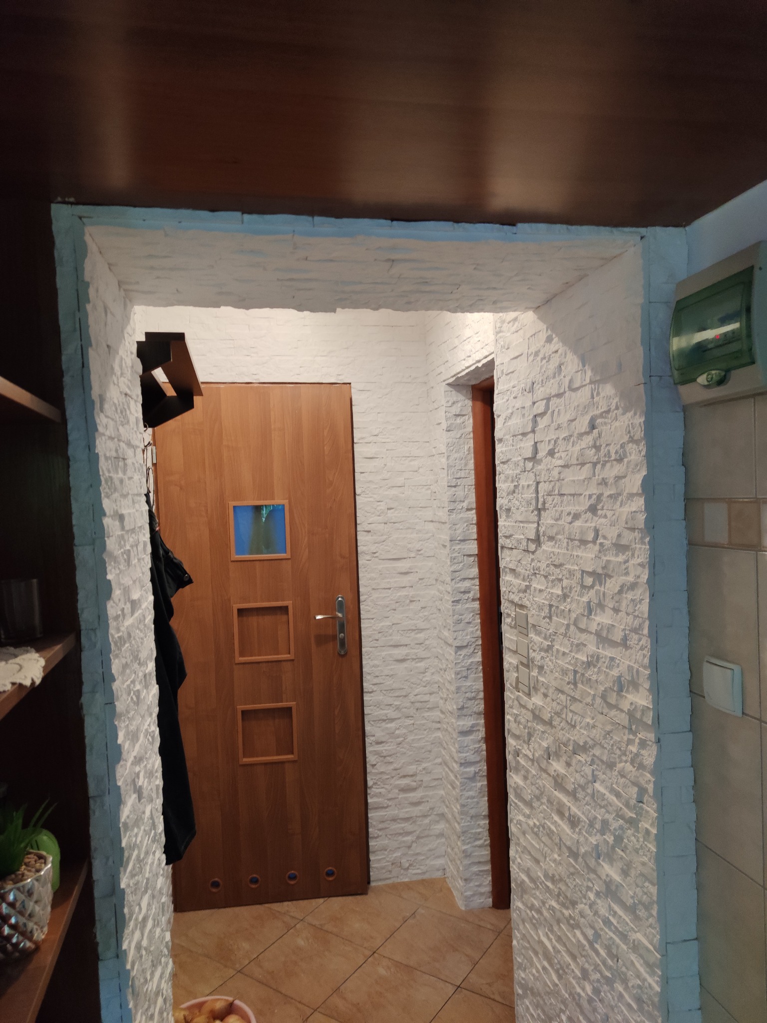 Korytarz z drewnianymi drzwiami z trzema kwadratowymi wgłębieniami, ściany wykończone białą cegłą dekoracyjną, widoczna szafka z półkami po lewej stronie.