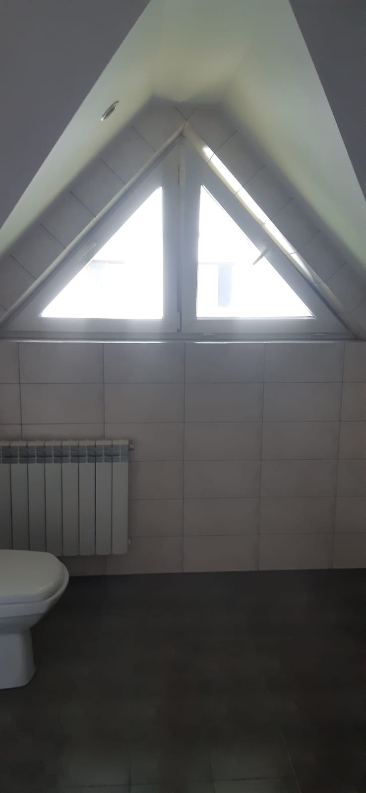 Wykończona łazienka na poddaszu z trójkątnym oknem, białe płytki na ścianach, ciemna podłoga, biały grzejnik i fragment toalety.