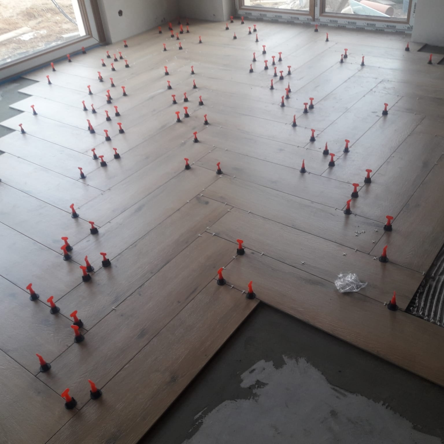 Układanie podłogi z płytek imitujących drewno w jodełkę, z użyciem czerwonych klipsów poziomujących, widoczne niedokończone fragmenty podłogi.