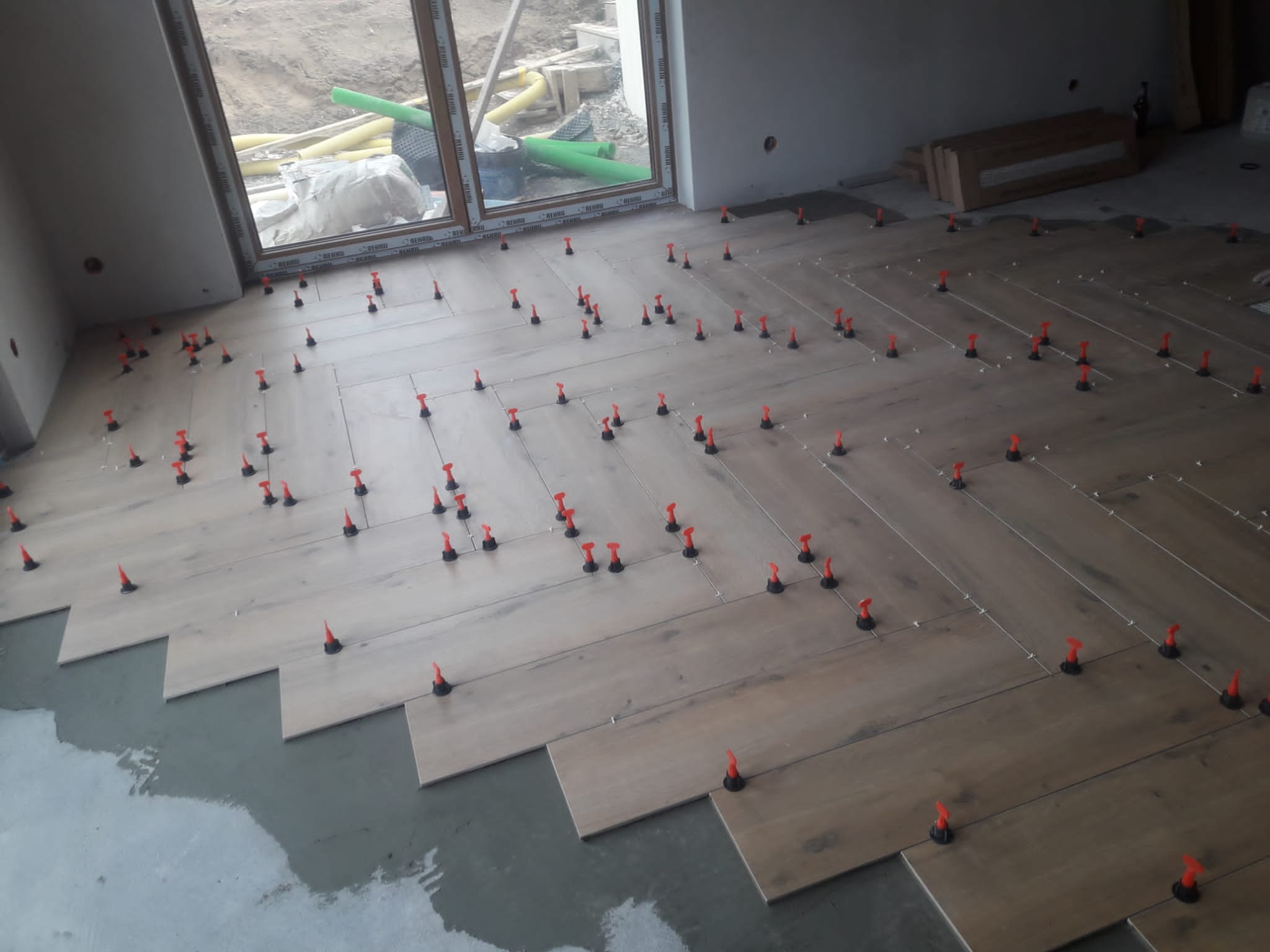 Układanie płytek podłogowych w jodełkę, z użyciem czerwonych klipsów poziomujących na świeżo wylanej zaprawie.