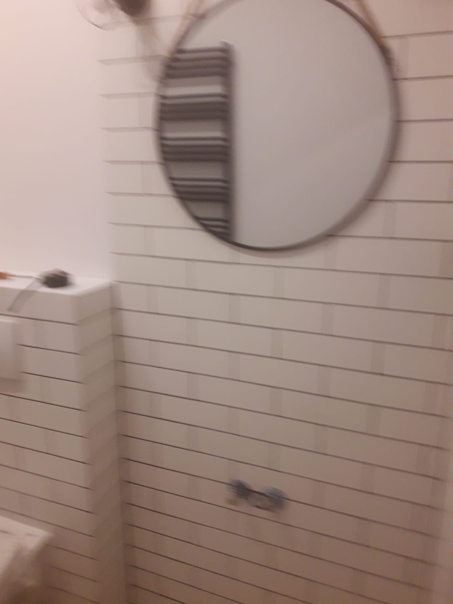 Rozmyte ujęcie fragmentu łazienki z białymi, prostokątnymi płytkami ułożonymi poziomo, okrągłym lustrem na ścianie i grzejnikiem drabinkowym odbijającym się w lustrze.